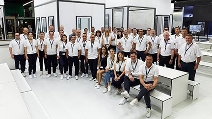 A group of people wearing white shirts and black pants are posing for a photo in a room with white walls and a white floor. Some are standing, while others are sitting on white benches.