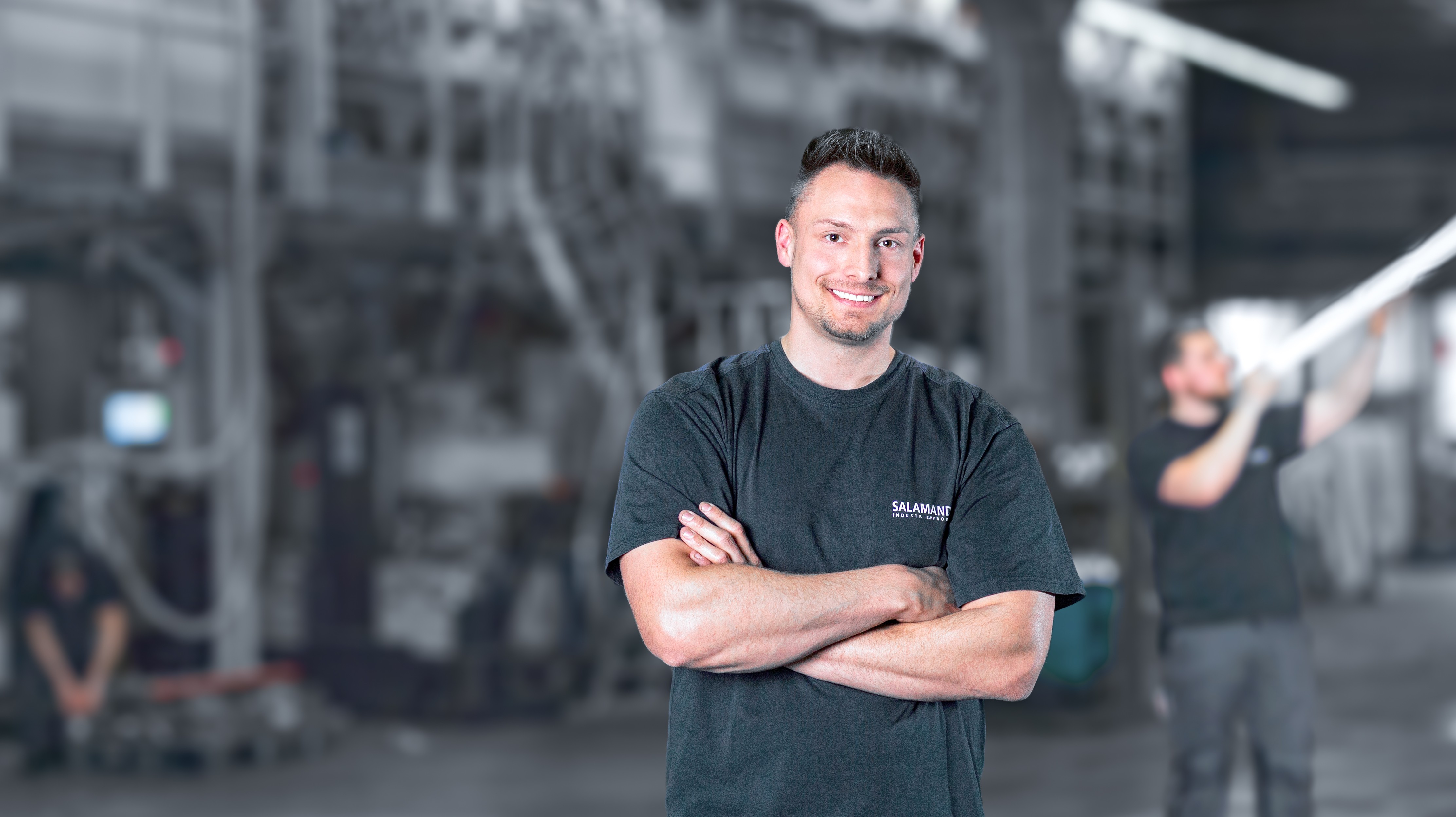 A man in a black t-shirt with the word "Salamand" on it stands with his arms crossed in front of a factory.
