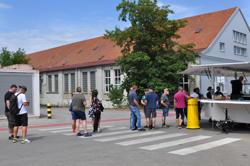 A group of people are standing in line outside a building with a red roof and a food truck.
