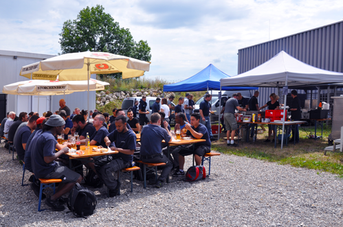 Um grupo de pessoas está sentado em mesas de piquenique sob guarda-sóis e tendas em um evento ao ar livre.