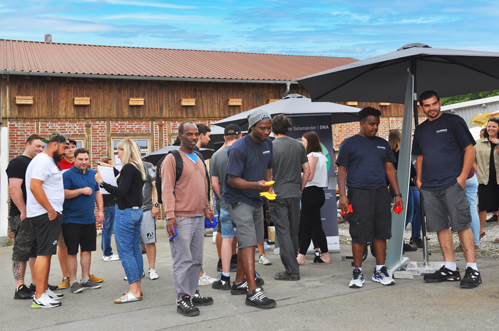A group of people are standing outside a building with a large umbrella providing shade. Some are wearing hats and sneakers, and one person is holding a yellow object.