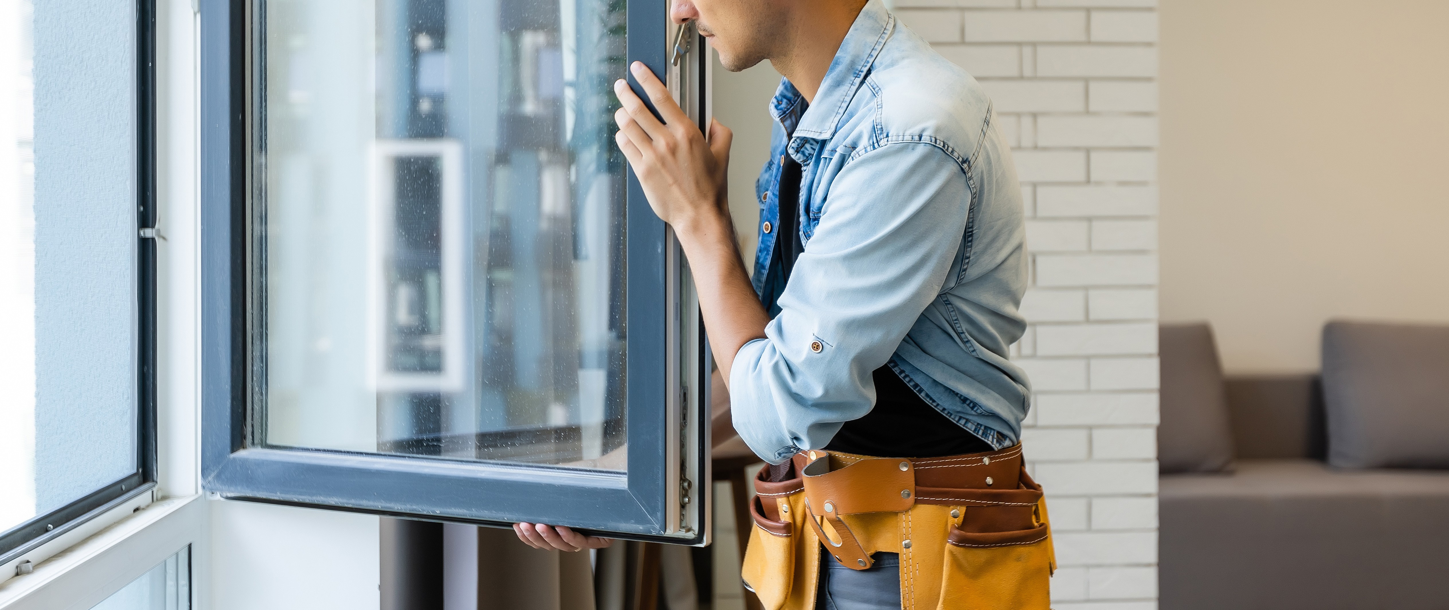 A man in a denim shirt and tool belt is opening a window in a room with a couch and brick wall.