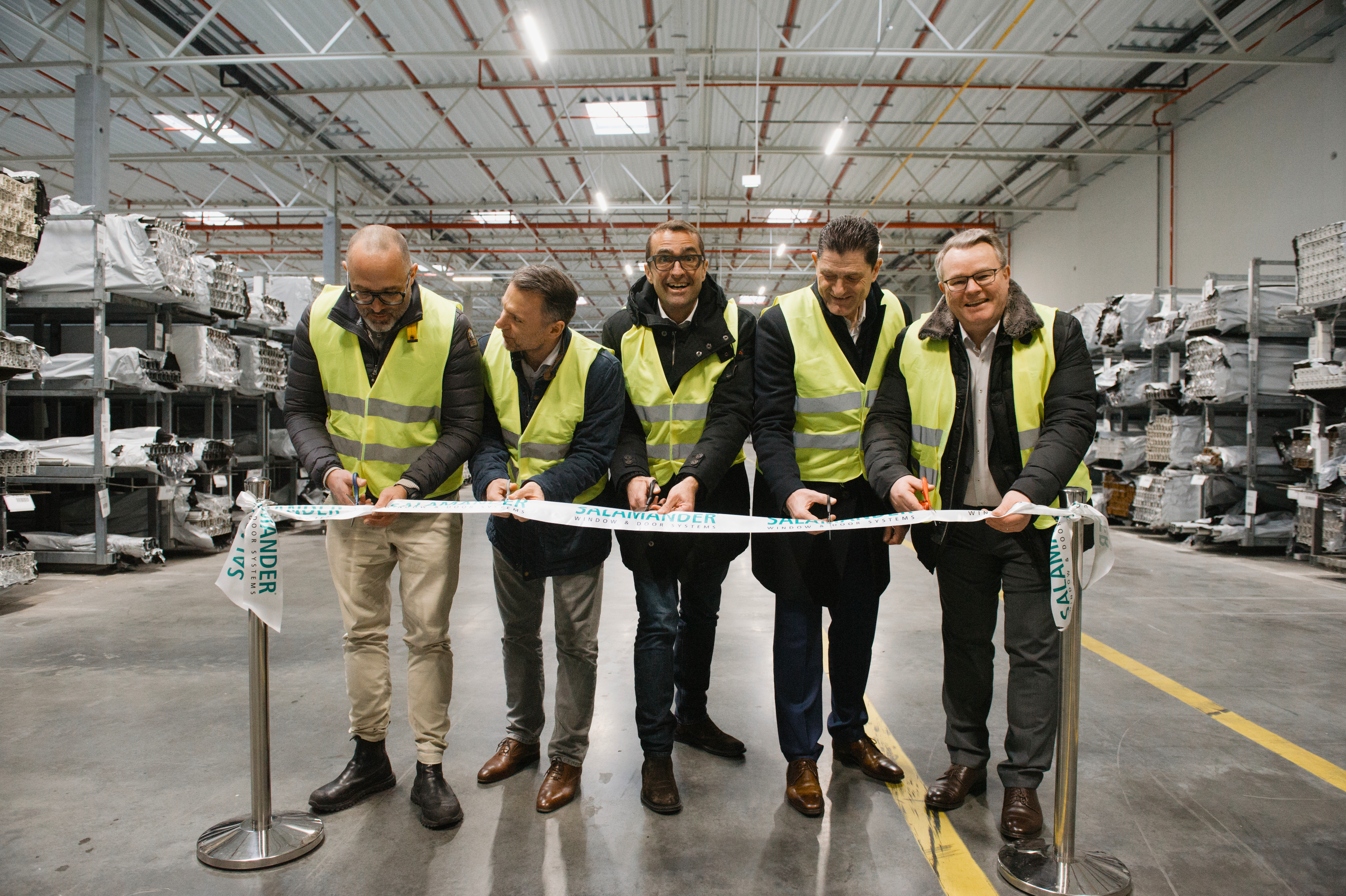 Cinco homens em coletes e jaquetas de segurança estão em um armazém, segurando uma fita com as palavras "Salamander Window & Door Systems" impressas nela. Eles estão sorrindo e parecem estar celebrando um evento. Atrás deles, prateleiras cheias de vários itens são visíveis, e o armazém tem um teto de estrutura metálica com luzes.