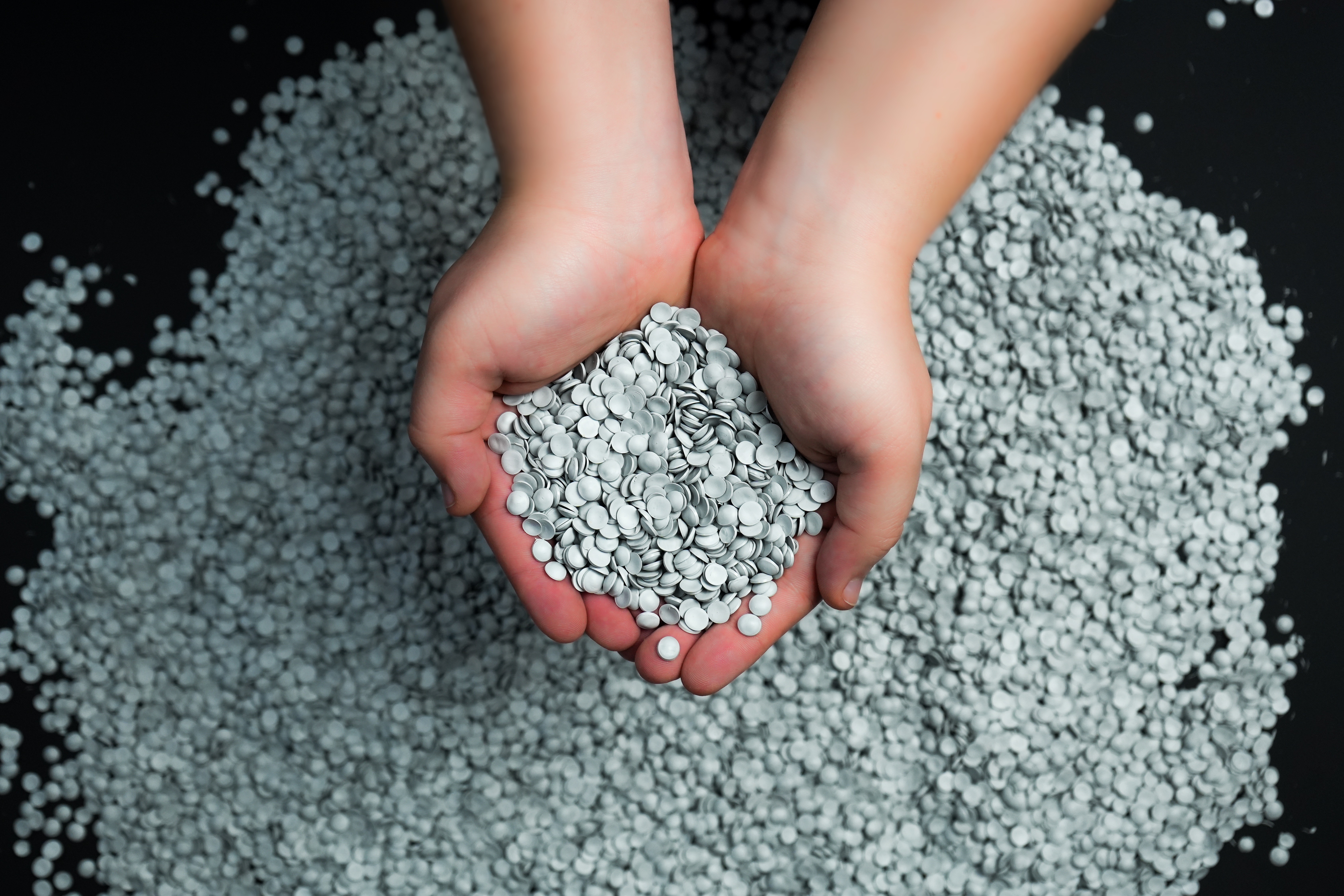 Un par de manos sosteniendo un puñado de pequeños gránulos de plástico redondos y grises, posiblemente de polipropileno, sobre un fondo negro.