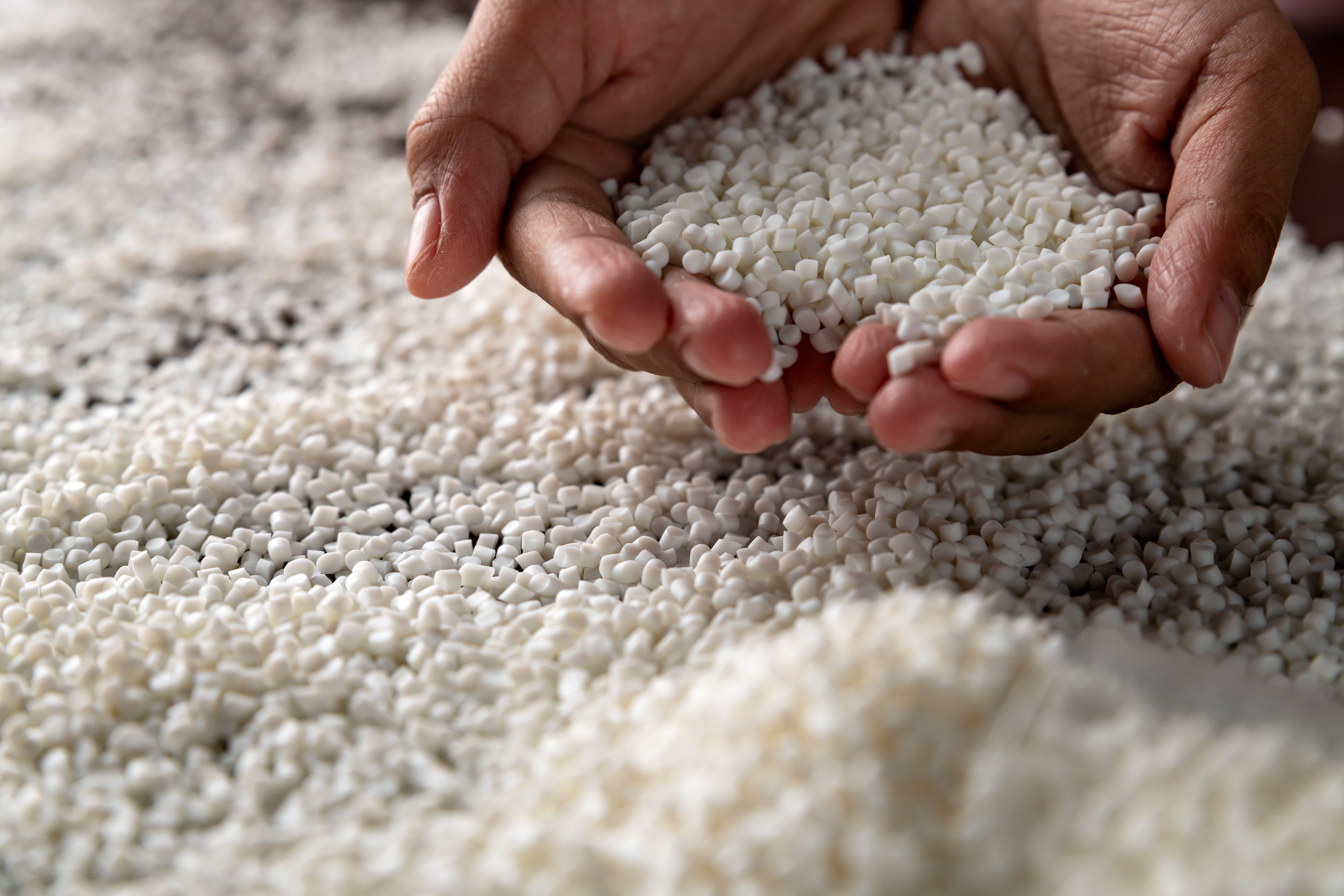 Manos sosteniendo una pila de gránulos de plástico blanco, posiblemente polipropileno, en un entorno de fábrica.