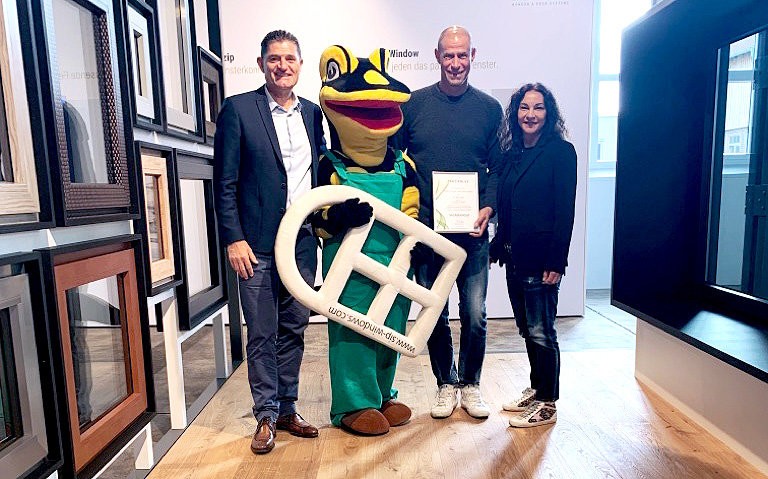Três pessoas e um mascote estão em uma sala, sorrindo e posando para uma foto. O mascote está segurando um certificado e uma grande letra H. O homem à esquerda está vestindo um terno e sapatos de couro, enquanto o homem à direita está vestindo uma camisa preta e tênis. A mulher à direita está vestindo um casaco preto e tênis. Atrás deles há molduras de fotos na parede.