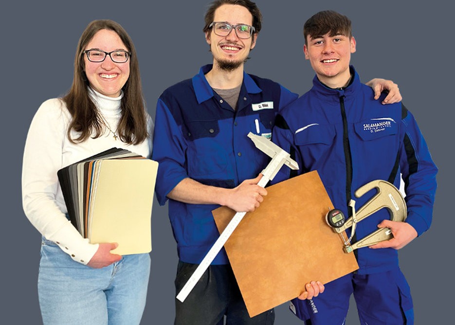 Three people in blue uniforms stand together, smiling. The woman on the left holds a folder, the man in the middle holds a ruler and a piece of wood, and the man on the right holds a measuring tool.