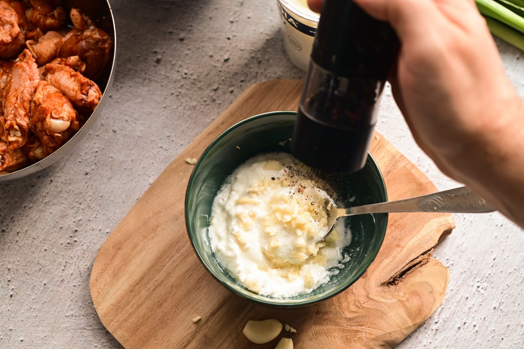 Ruka dochucující jogurtovo-česnekovou směs v zelené misce na dřevěném prkénku, vedle naložené kuřecí maso. Recept připravil Vojta Poštulka.