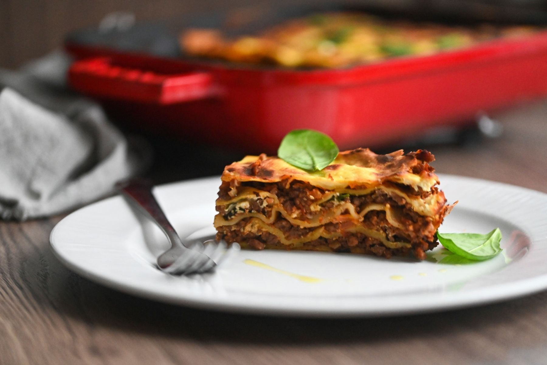 Porce lasagní na bílém talíři ozdobená bazalkou, vedle leží vidlička. V pozadí je červená zapékací mísa s dalšími lasagněmi. Recept připravil Vojta Poštulka.