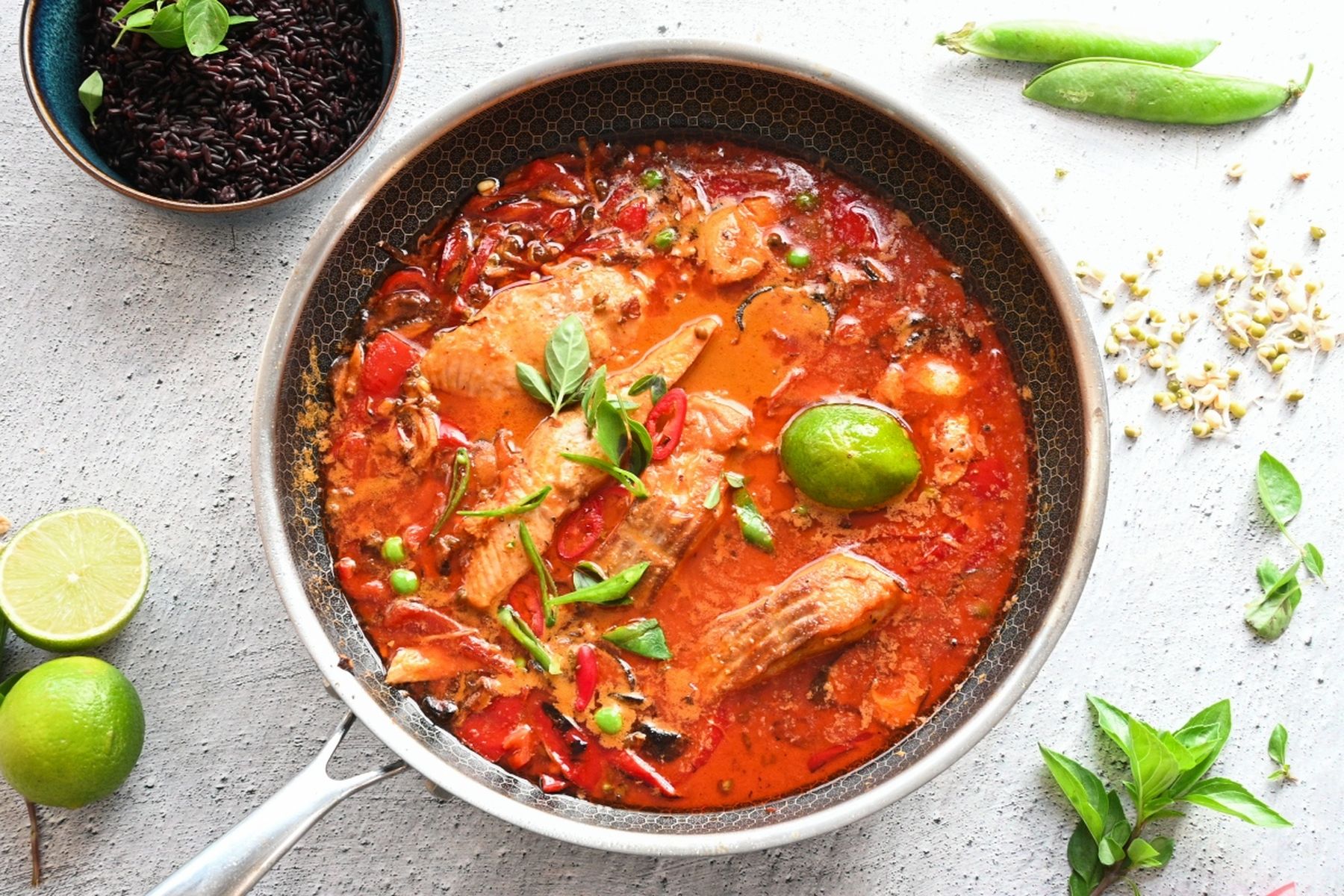 Pánev s pikantním červeným curry s filety z lososa, ozdobená čerstvými bylinkami a limetkou. V misce černá rýže a kolem čerstvé suroviny. Recept připravil Vojta Poštulka.
