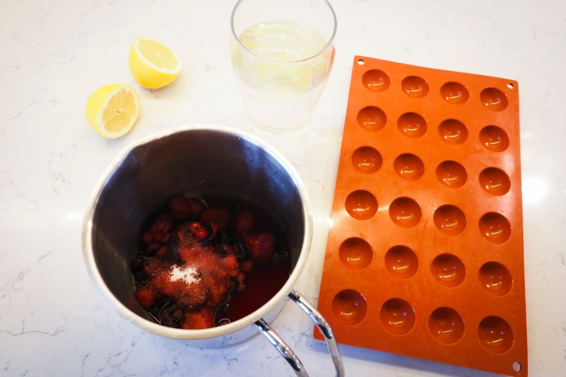 Kovový hrnec s uvařenou ovocnou směsí, silikonová forma, sklenice s rozpuštěnou želatinou a dvě poloviny citronu na bílé pracovní desce.