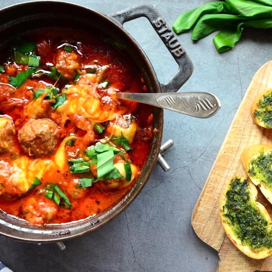 Hrnec s masovými kuličkami v rajčatové omáčce posypanými čerstvou bazalkou, vedle dřevěné desky s plátky chleba s pestem a svazkem medvědího česneku.