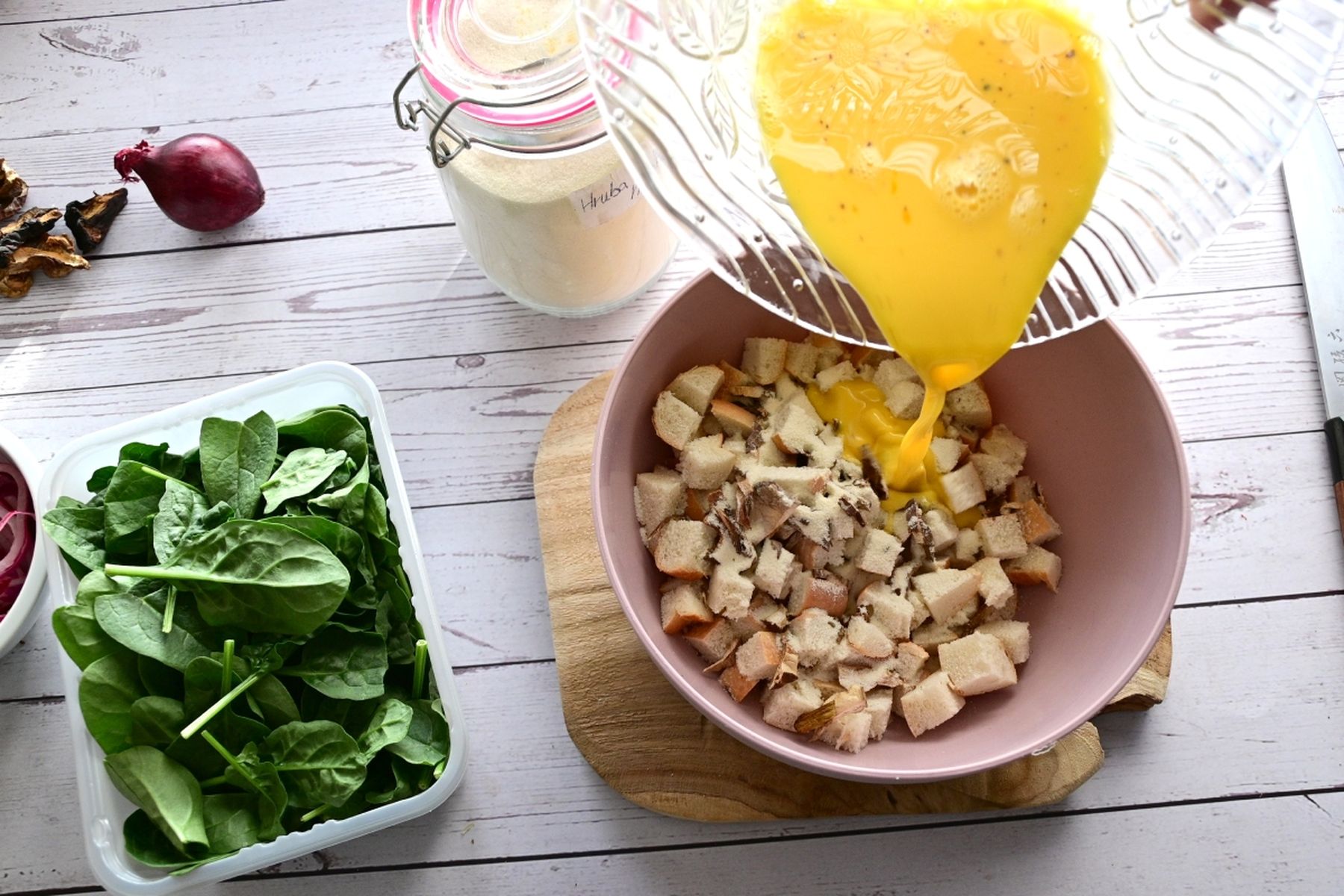 Vaječná směs nalévaná do mísy s kostičkami chleba, kolem špenát, sklenice s moukou, červená cibule a nůž na dřevěném stole. Recept připravil Vojta Poštulka.