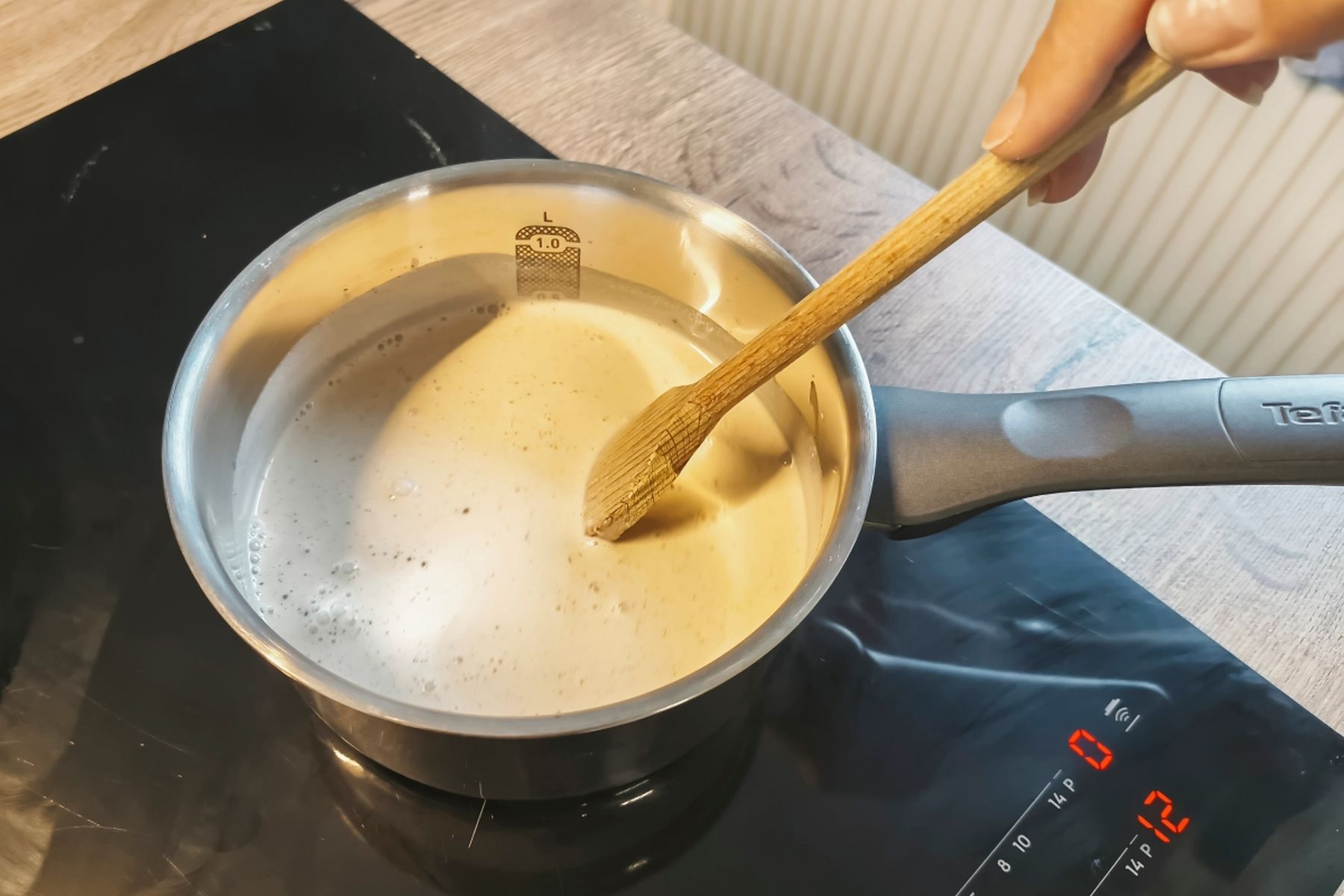 Osoba míchá krémovou tekutinu v malém rendlíku dřevěnou stěrkou na elektrické varné desce. Recept připravily Ségry v kuchyni.