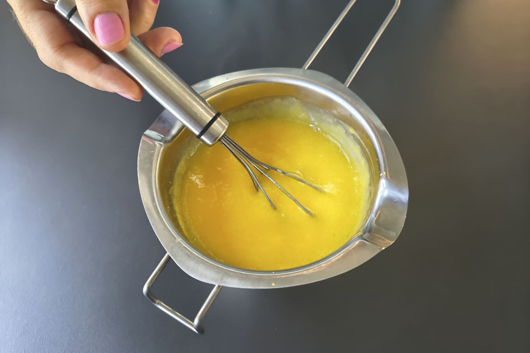 Ruka šlehá žlutou směs v nerezové míse na tmavé pracovní ploše. Recept připravily Ségry v kuchyni.