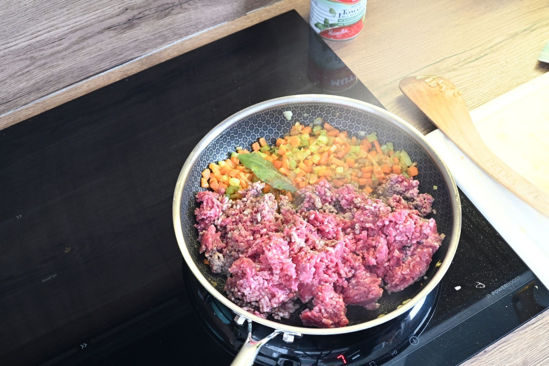 Mleté maso připravované na pánvi s nakrájenou mrkví, cibulí a bobkovým listem na sporáku, vedle leží dřevěná vařečka. Recept připravil Vojta Poštulka.