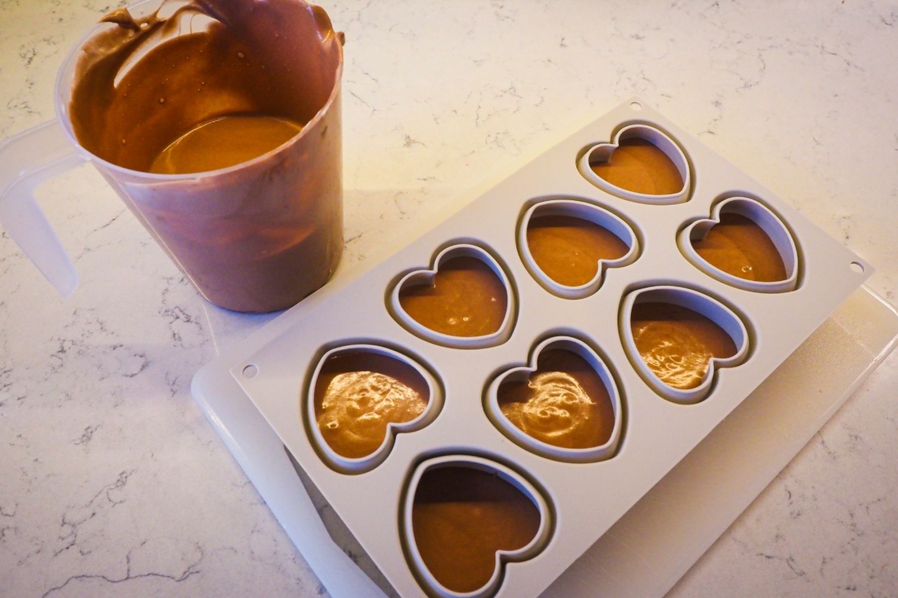 Silikonová forma ve tvaru srdce naplněná čokoládovou pěnou vedle nádoby s další pěnou na mramorové pracovní desce.