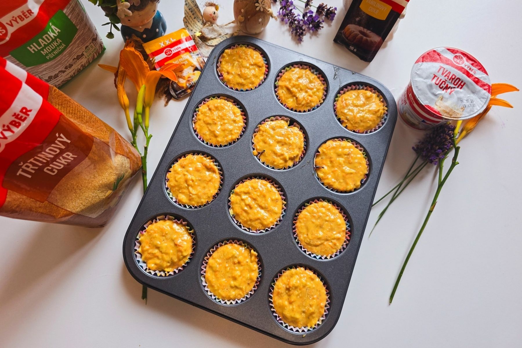 Plech na muffiny s neupečeným mrkvovo-tvarohovým těstem v papírových košíčcích, obklopený ingrediencemi na pečení a květinami na bílém stole.