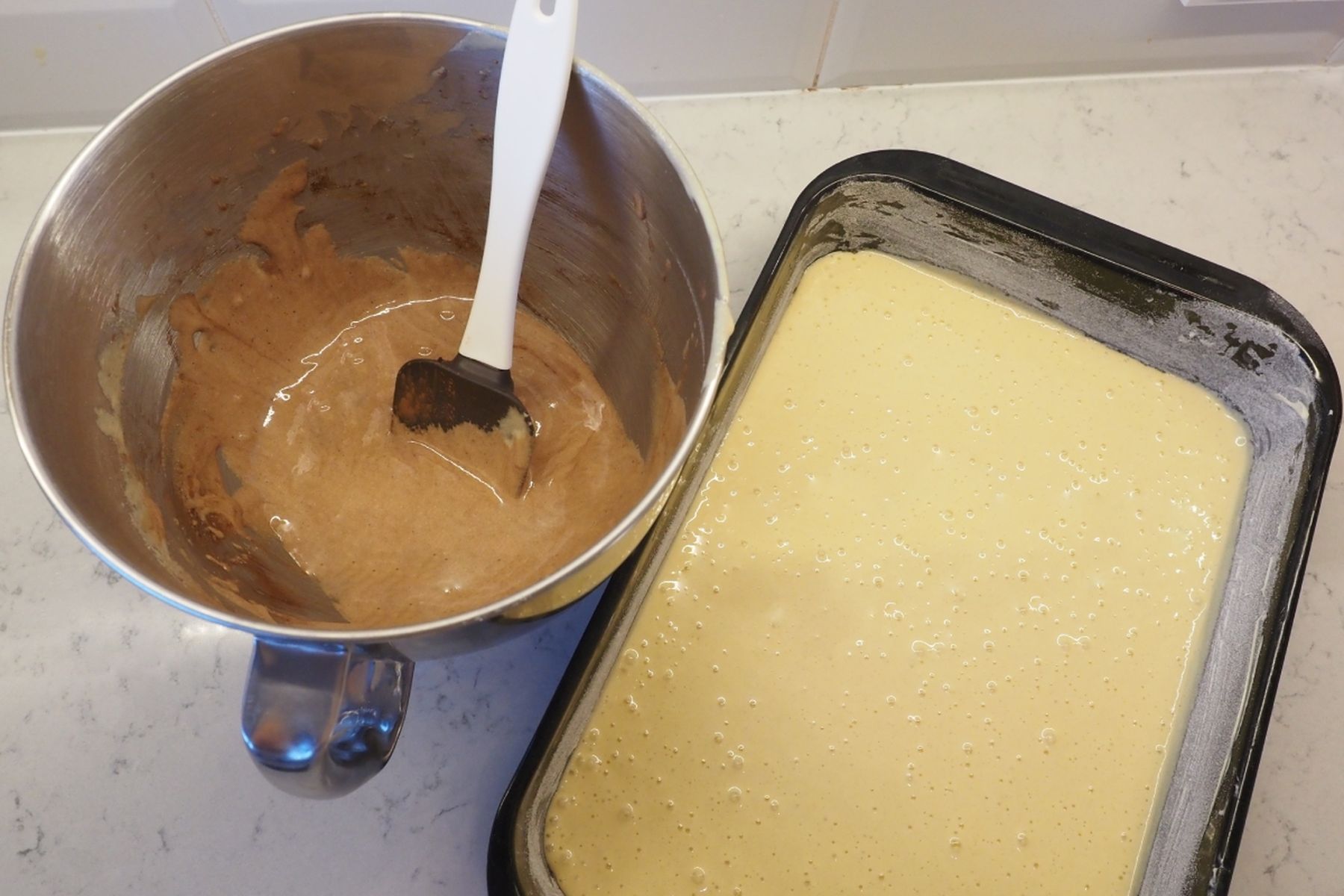 Kovová mísa s čokoládovým těstem a stěrkou vedle vymazaného obdélníkového pekáče naplněného světlým těstem na pracovní desce.