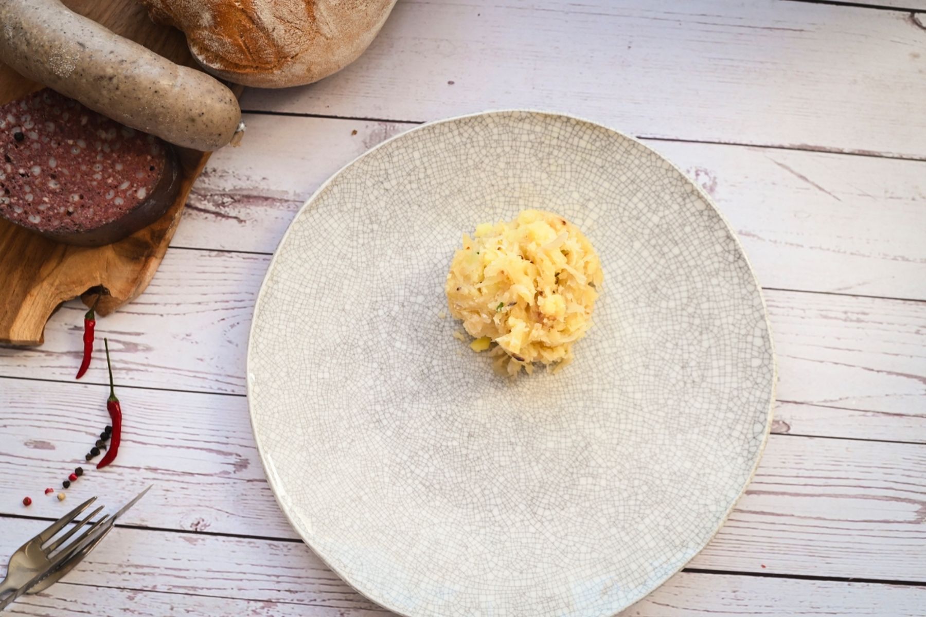 Kopec bramborové kaše na kulatém, strukturovaném talíři, v blízkosti dřevěného prkénka s jitrnicí, prejtem a chlebem.