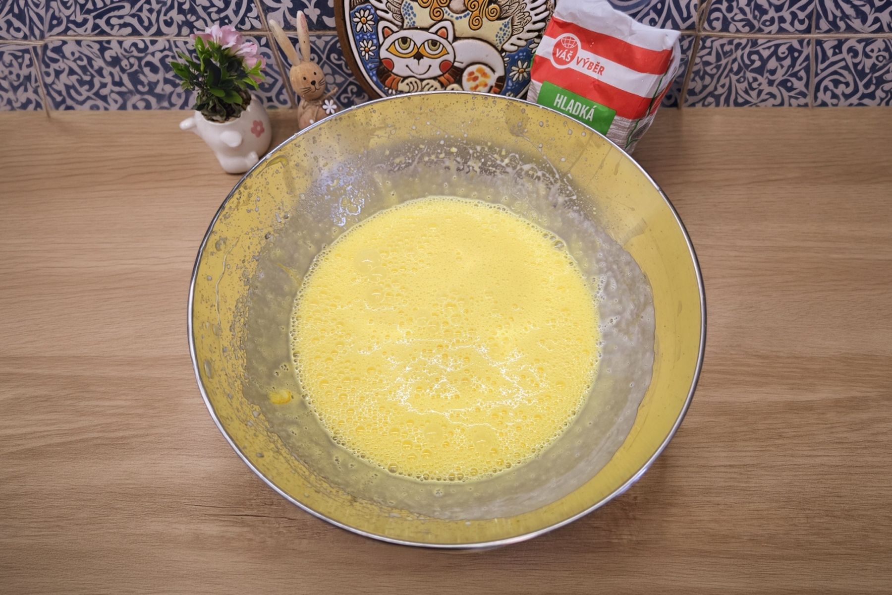 Mísa s těstem na dřevěné pracovní desce, kolem kuchyňské dekorace, rostlina a ozdobný talíř. Recept připravil Táta v akci.