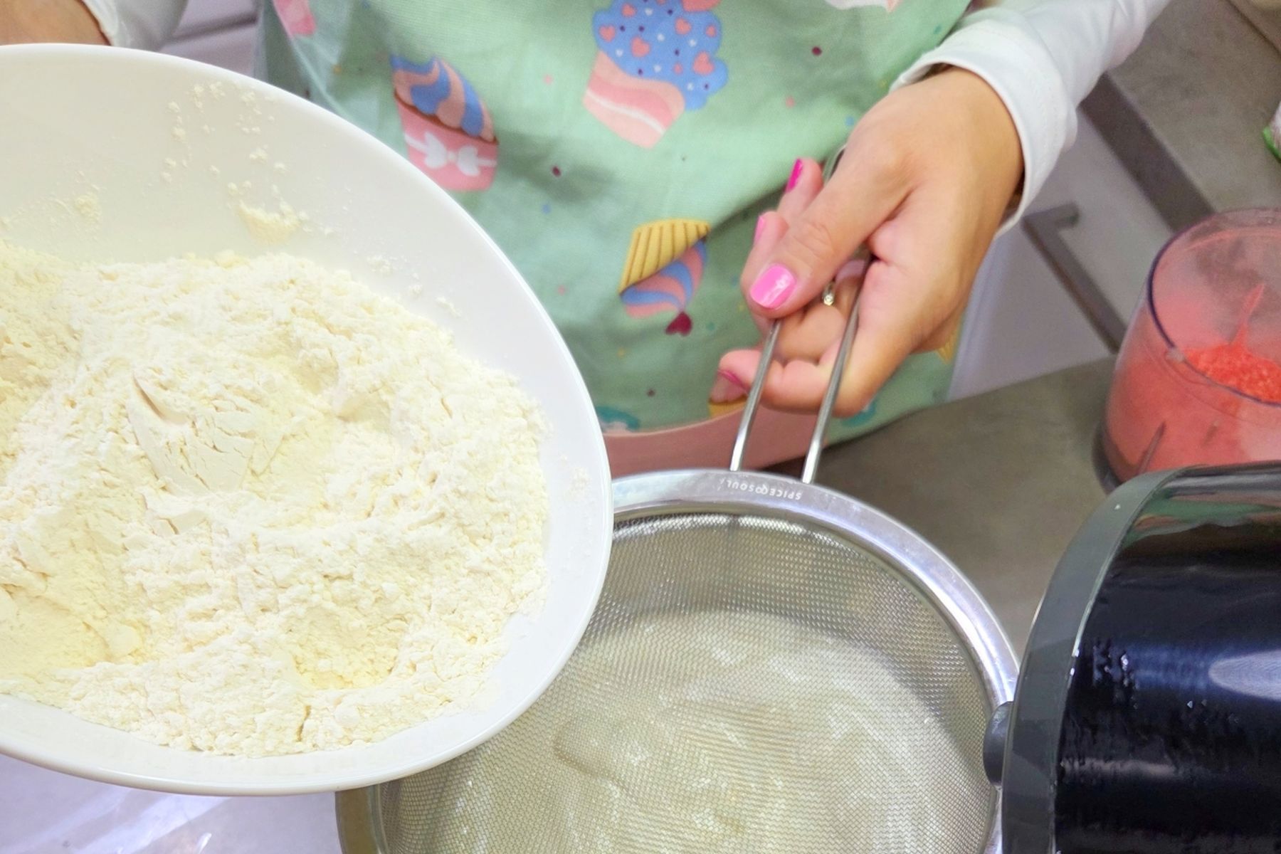Osoba, která v kuchyni prosívá mouku do mísy, drží v ruce síto a mísu a má na sobě zástěru s motivy cupcakeů.