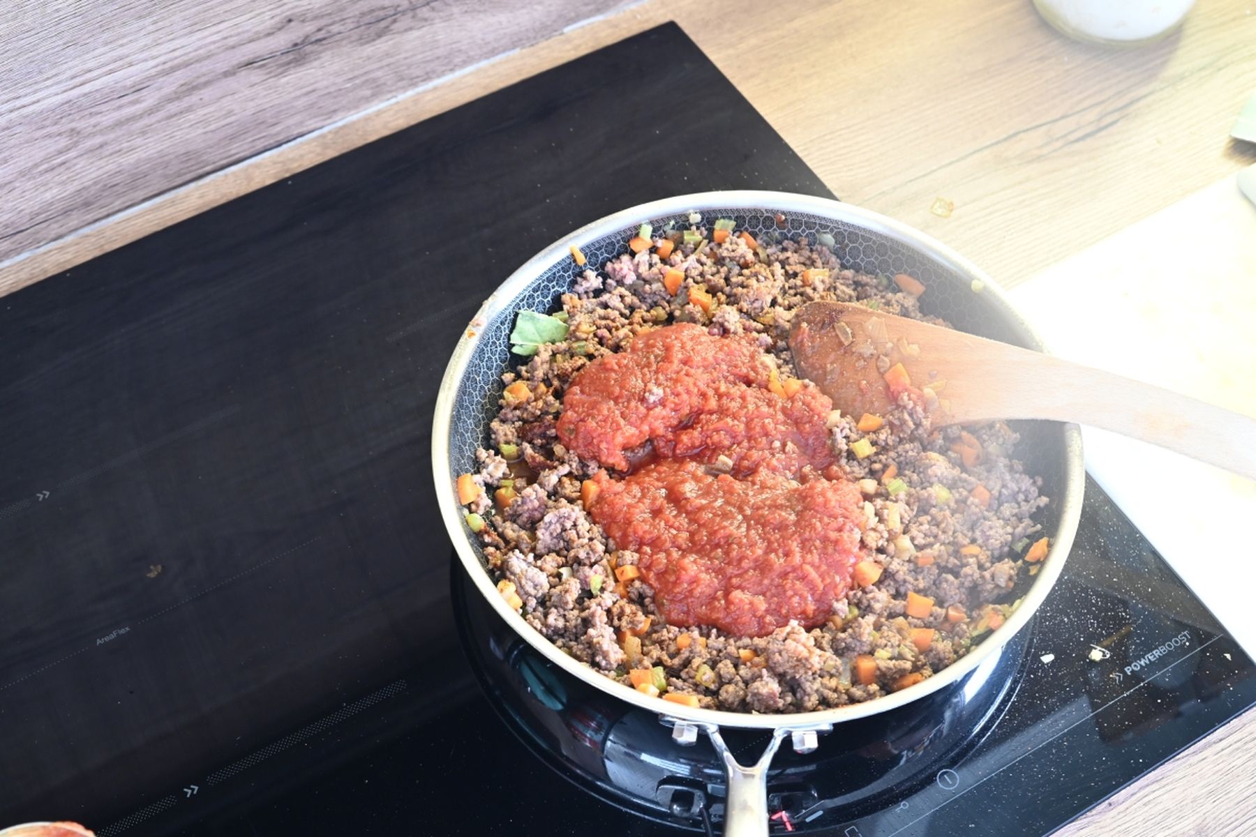 Pánev s mletým masem, nakrájenou zeleninou a rajčatovou omáčkou, která se míchá dřevěnou lžící na sporáku v kuchyni.