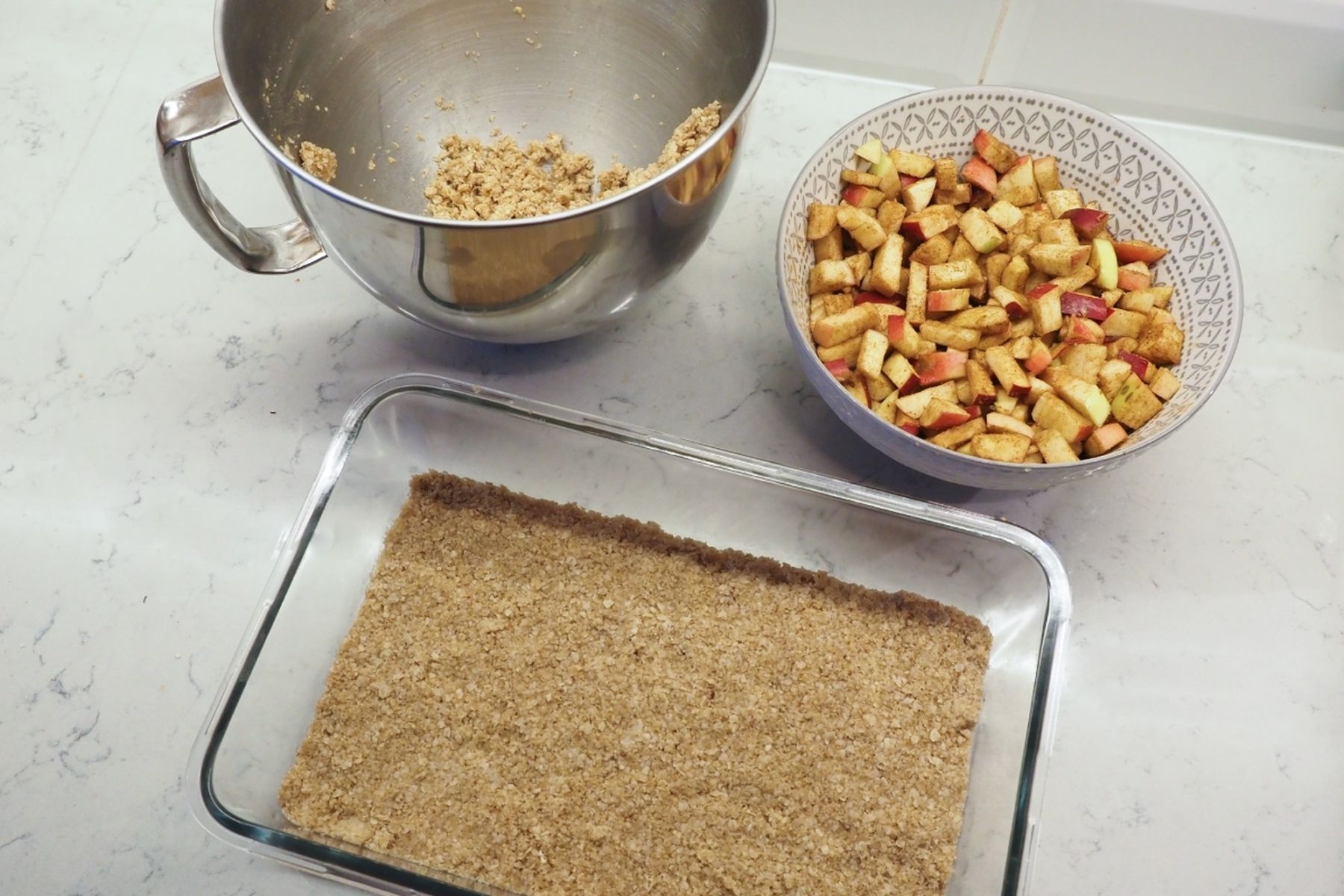 Pečicí sestava s kovovou miskou s drobenkou, miskou s nakrájenými jablky a skleněnou formou se základem drobenky na mramorovém povrchu, podle receptu Kitchen Affair na Jablečné řezy z webu hypermarketu Globus.