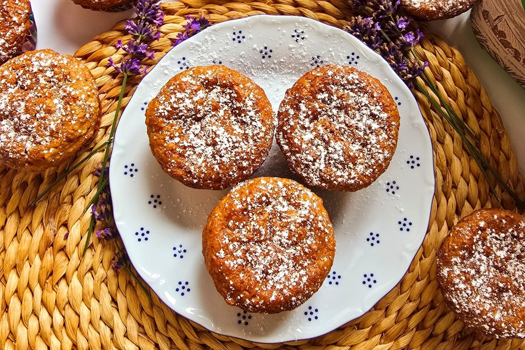 Tři muffiny posypané moučkovým cukrem na bílém talíři s proutěnou podložkou a větvičkami levandule pod nimi.