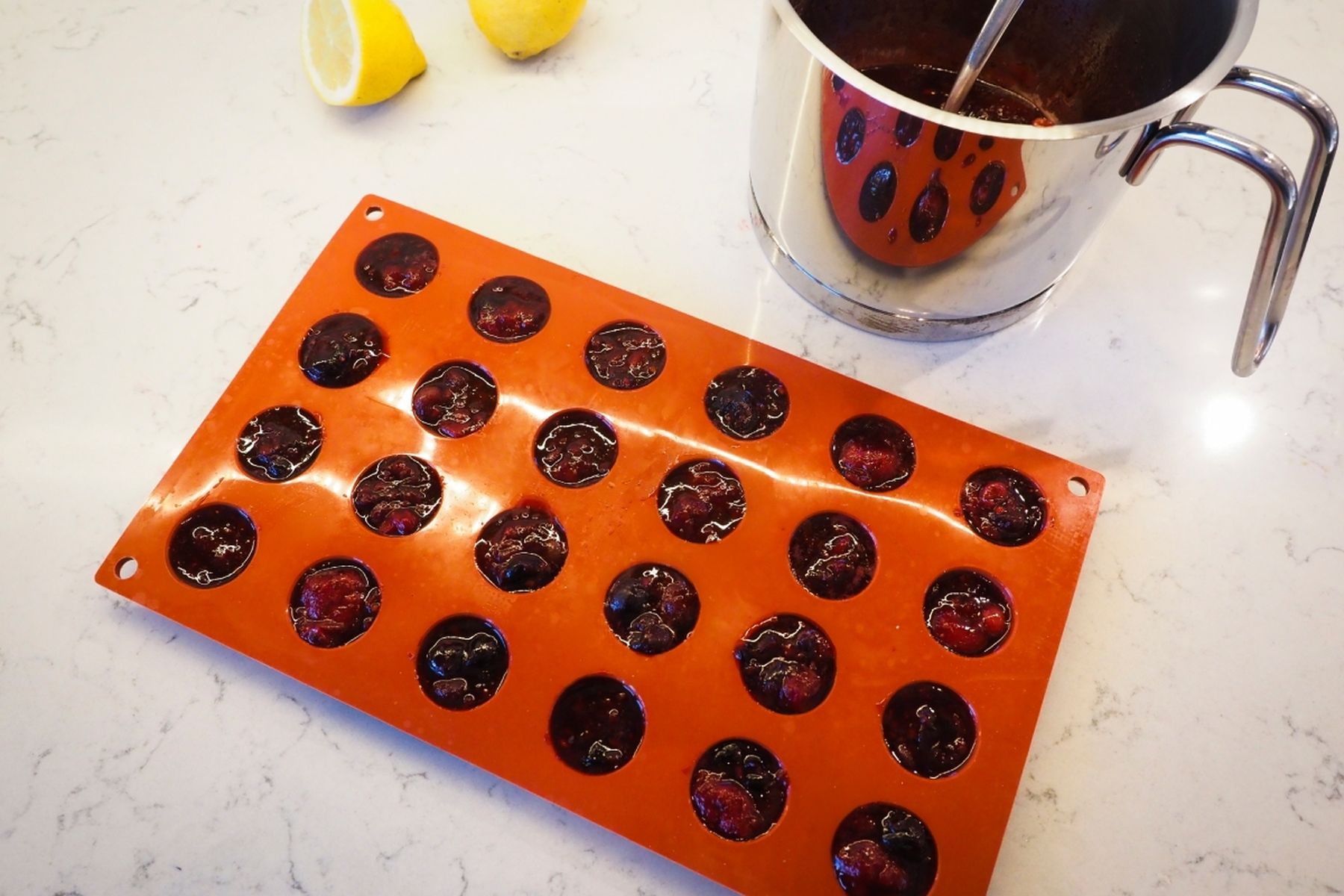 Ovocná směs v oranžových silikonových formičkách vedle kovového hrnce na bílé pracovní desce, příprava ingrediencí.