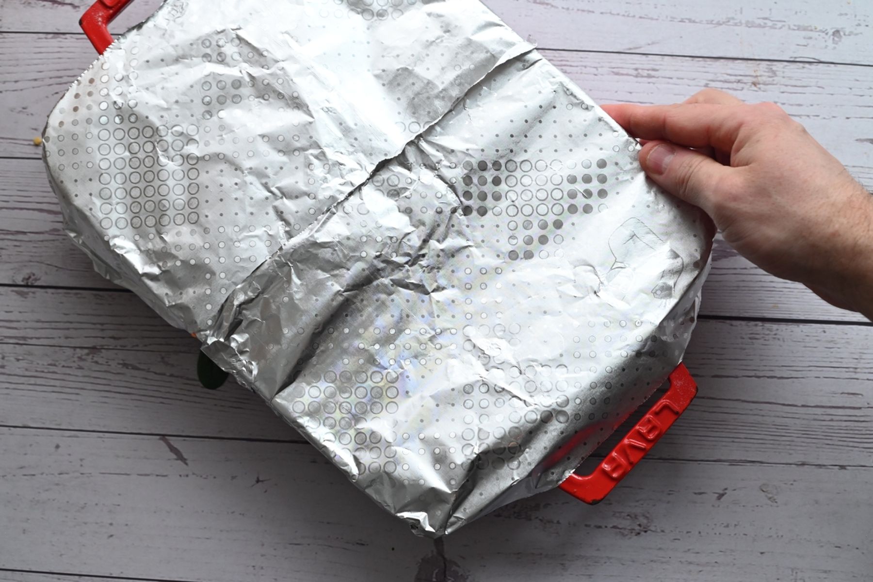 Ruka držící obdélníkovou zapékací mísu s červenými úchyty zakrytou alobalem na dřevěném povrchu.