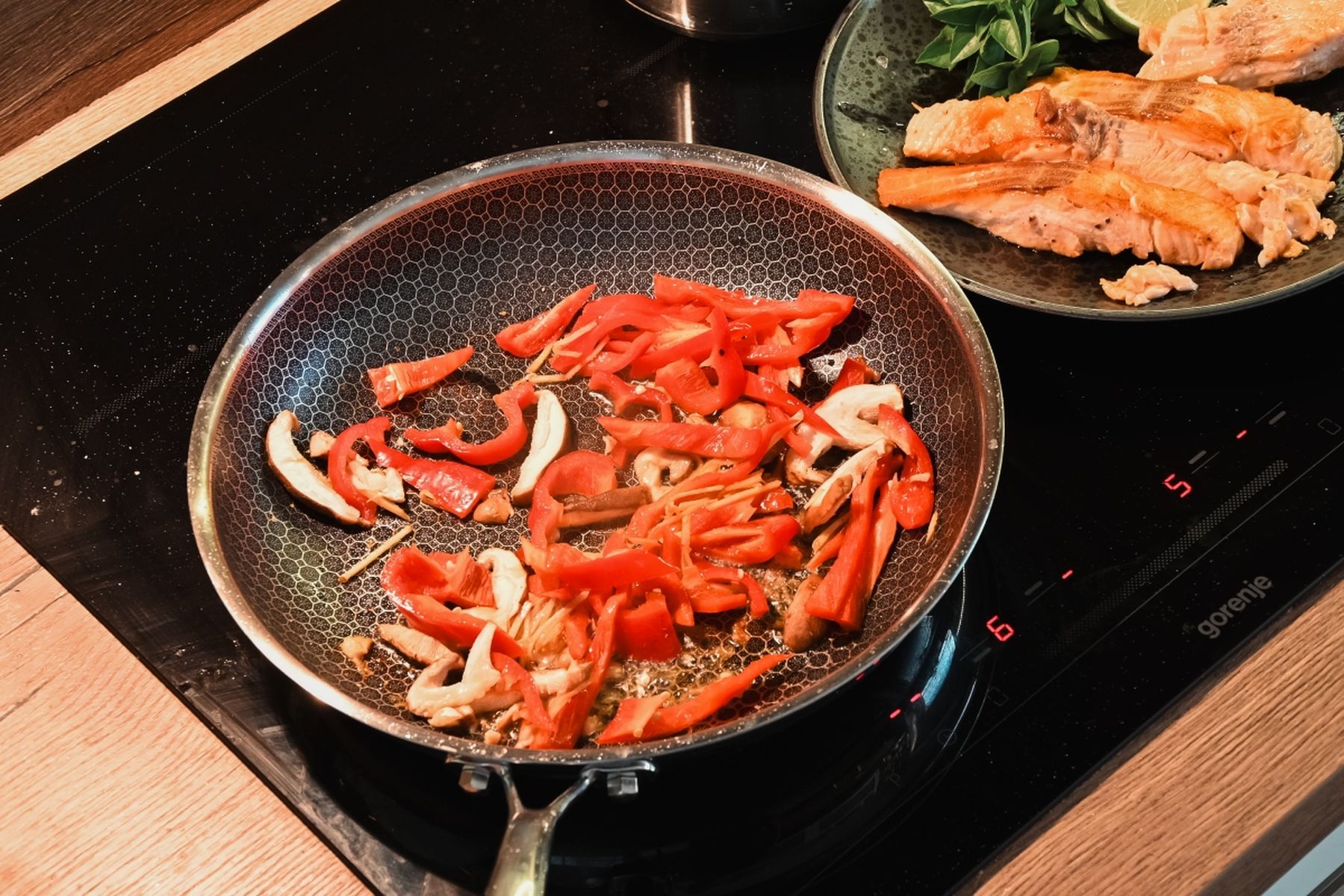 Nakrájené červené papriky a žampiony se restují na pánvi, vedle na talíři je připravený losos a bylinky na černé varné desce. Recept připravil Vojta Poštulka.