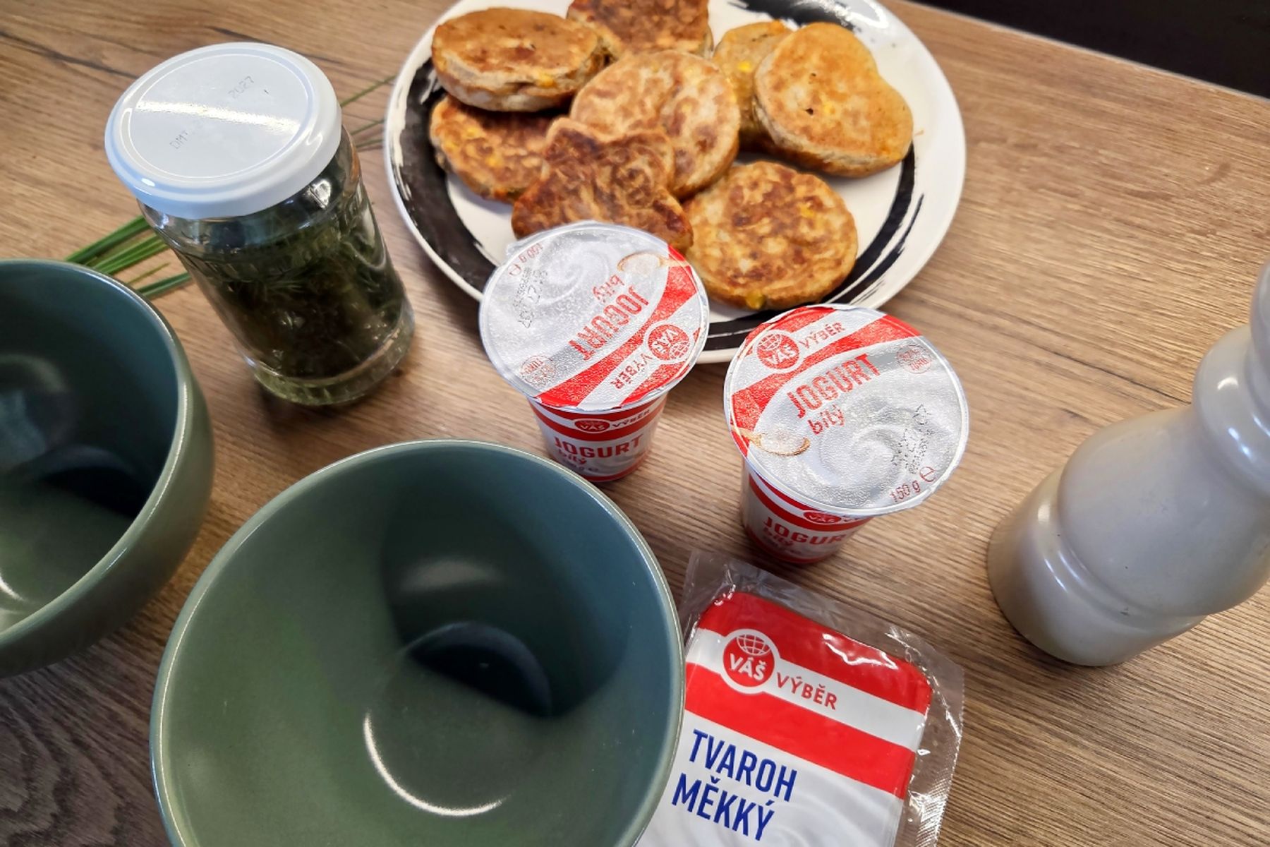 Stůl se dvěma zelenými miskami, sklenicí s koprem, kelímky jogurtu, tvarohem a talířem křupavých slaných lívanců. Recept připravily Ségry v kuchyni.