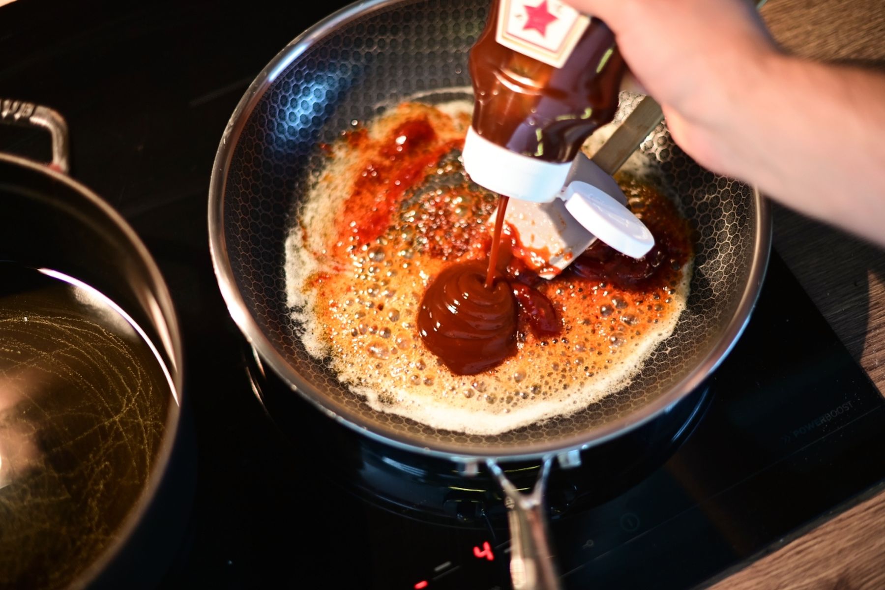 Kuchař nalévá BBQ omáčku do pánve na sporáku, kde se mísí s probublávajícím základem. Recept připravil Vojta Poštulka.