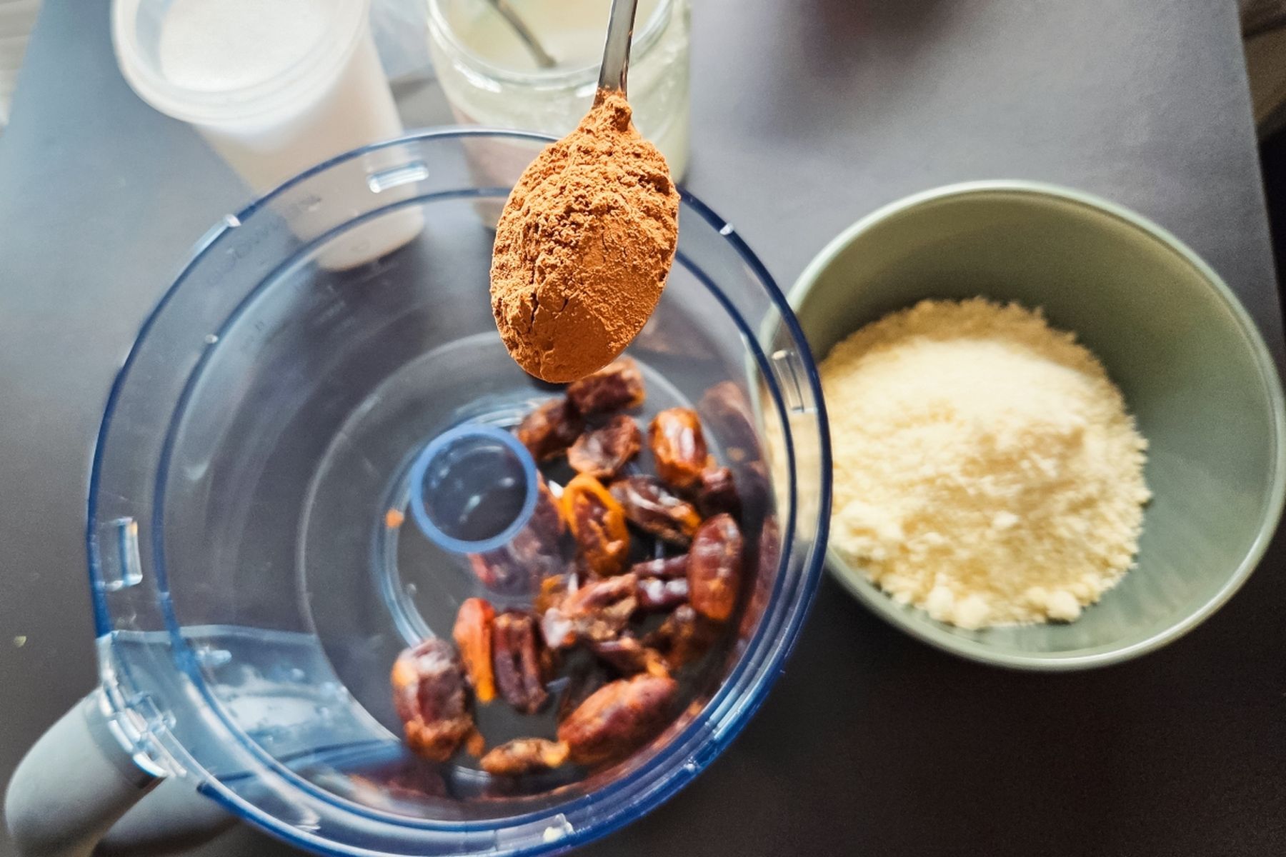 Lžíce kakaového prášku nad kuchyňským robotem s datlemi, vedle miska s mandlovou moukou a sklenice s dalšími surovinami. Recept připravily Ségry v kuchyni.