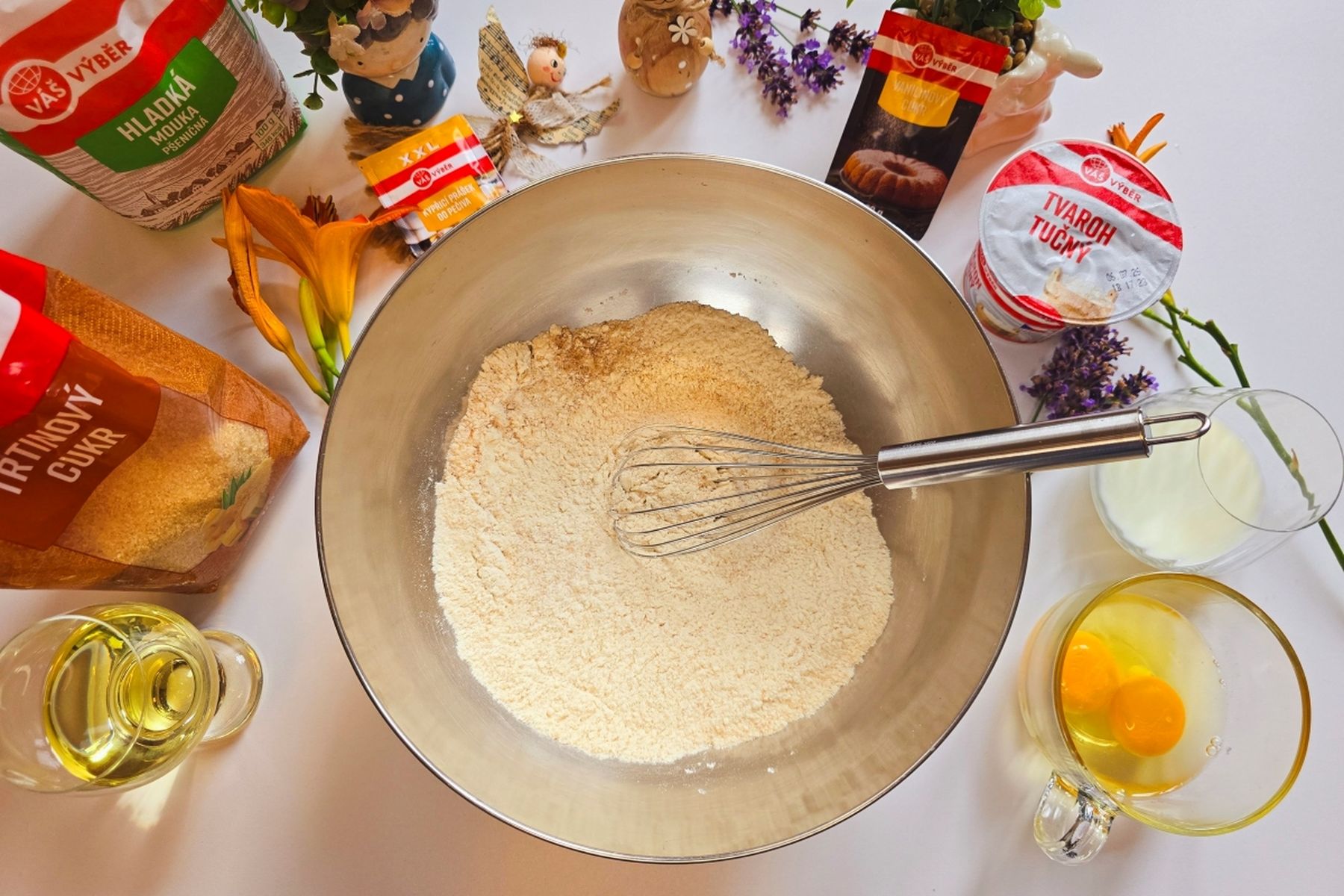 Mísa s moukou, dalšími suchými surovinami a metličkou, obklopená surovinami na pečení: vejci, olejem, mlékem, sáčky s cukrem a dalšími ingrediencemi, na bílém povrchu.