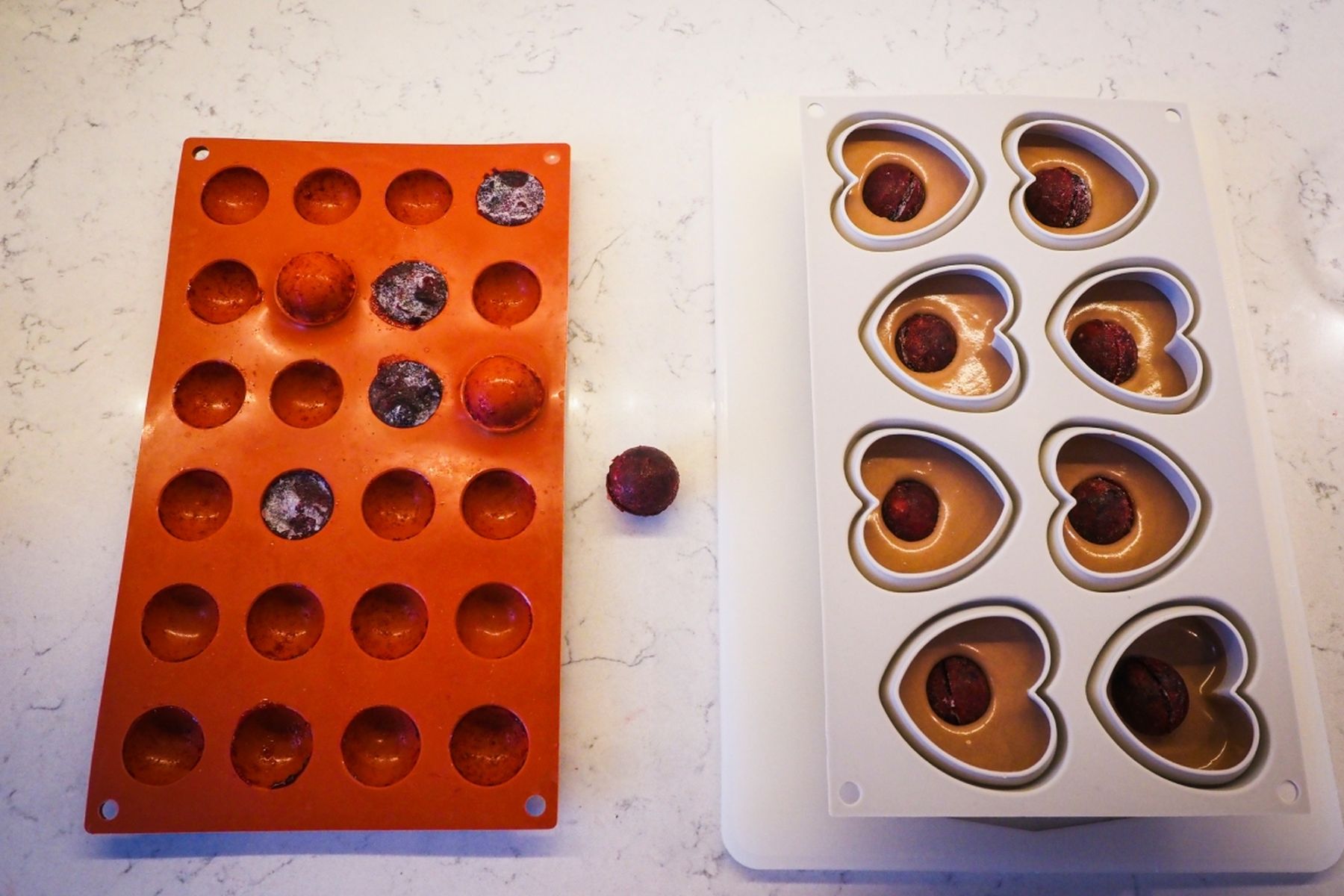 Silikonové formy na mramorové desce: červená forma na koule a několika ovocnými koulemi, bílá forma ve tvaru srdce částečně naplněná čokoládovou pěnou a ovocnými koulemi.