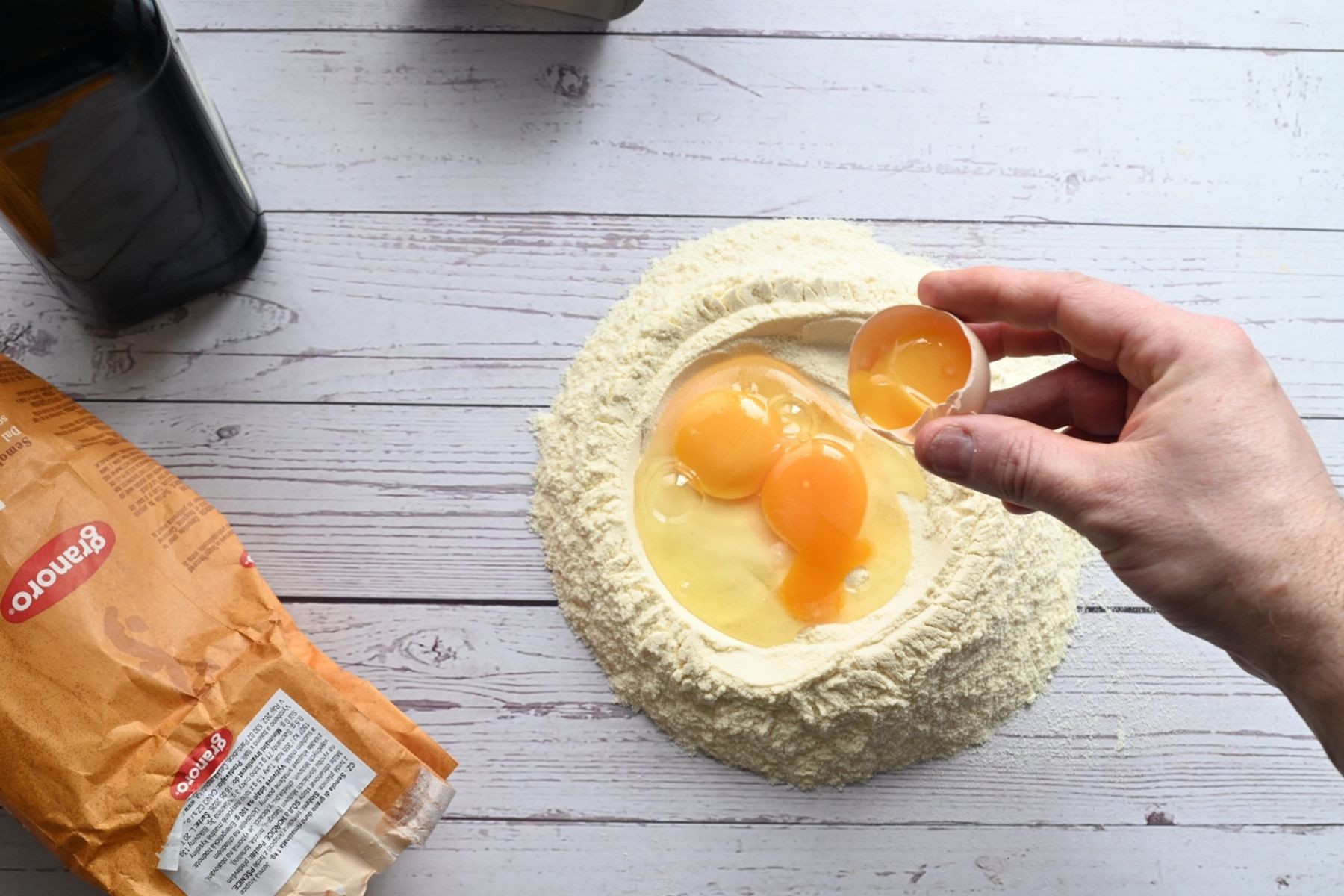 Osoba rozklepává vejce do důlku z mouky na dřevěném povrchu, kolem je láhev olivového oleje a sáček s moukou. Recept na lasagne připravil Vojta Poštulka.