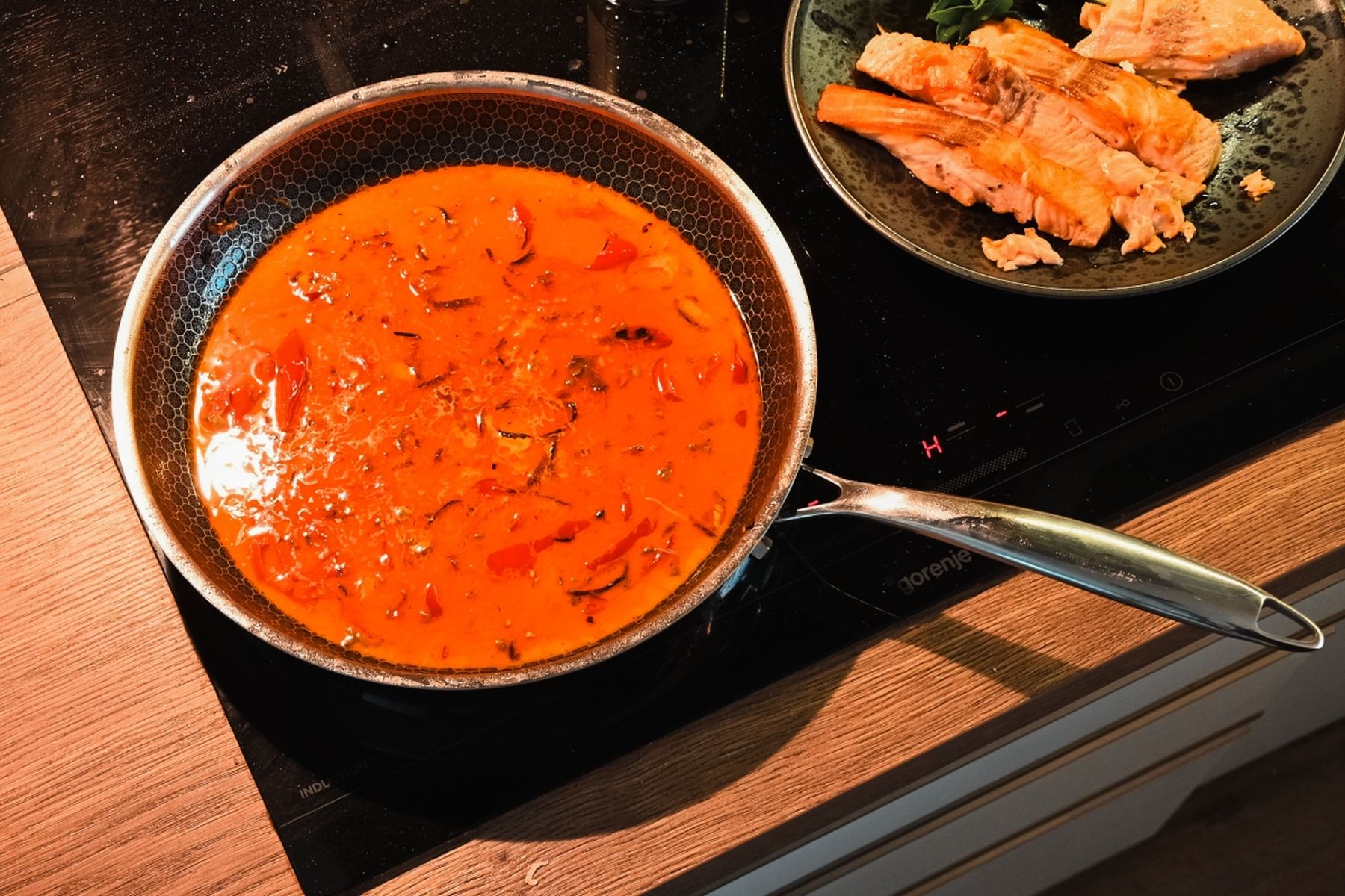 Pánev s červenou curry omáčkou se vaří na sporáku, vedle talíř s kousky připraveného lososa ozdobenými zelenými bylinkami. Recept připravil Vojta Poštulka.