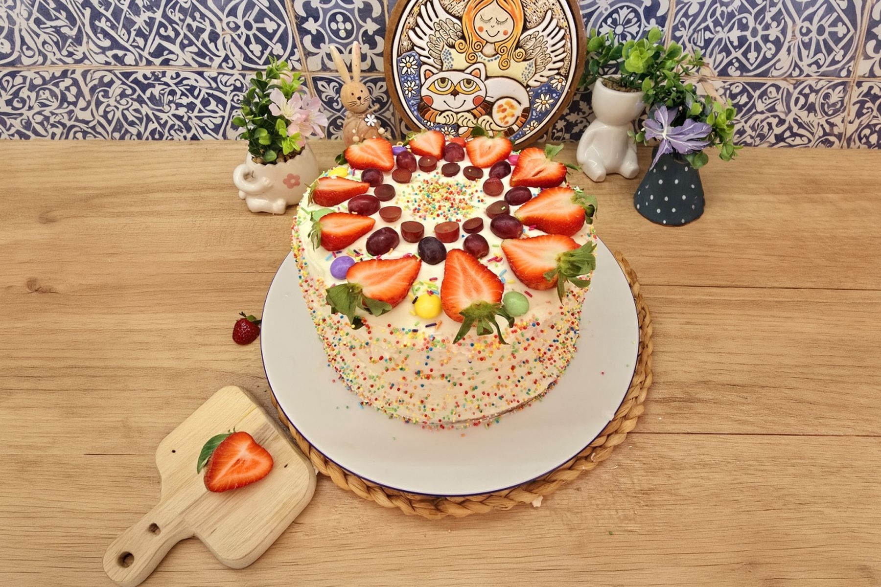 Barevný dort ozdobený jahodami a hroznovým vínem na dřevěném stole, obklopený dekorativními rostlinami a keramickým talířem. Recept připravil Táta v akci.
