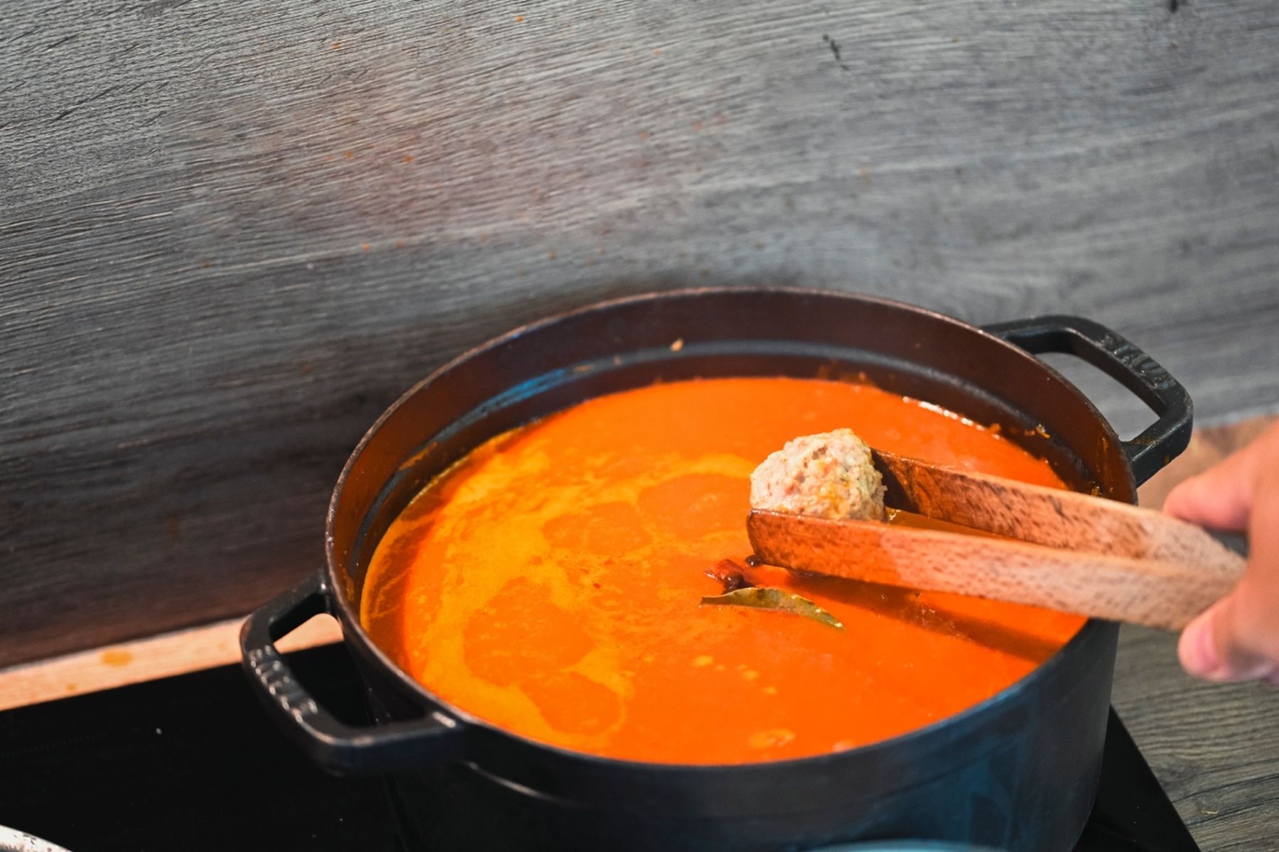Osoba drží masovou kuličku kleštěmi nad hrncem s hustou rajčatovou omáčkou na sporáku. Recept připravil Vojta Poštulka.
