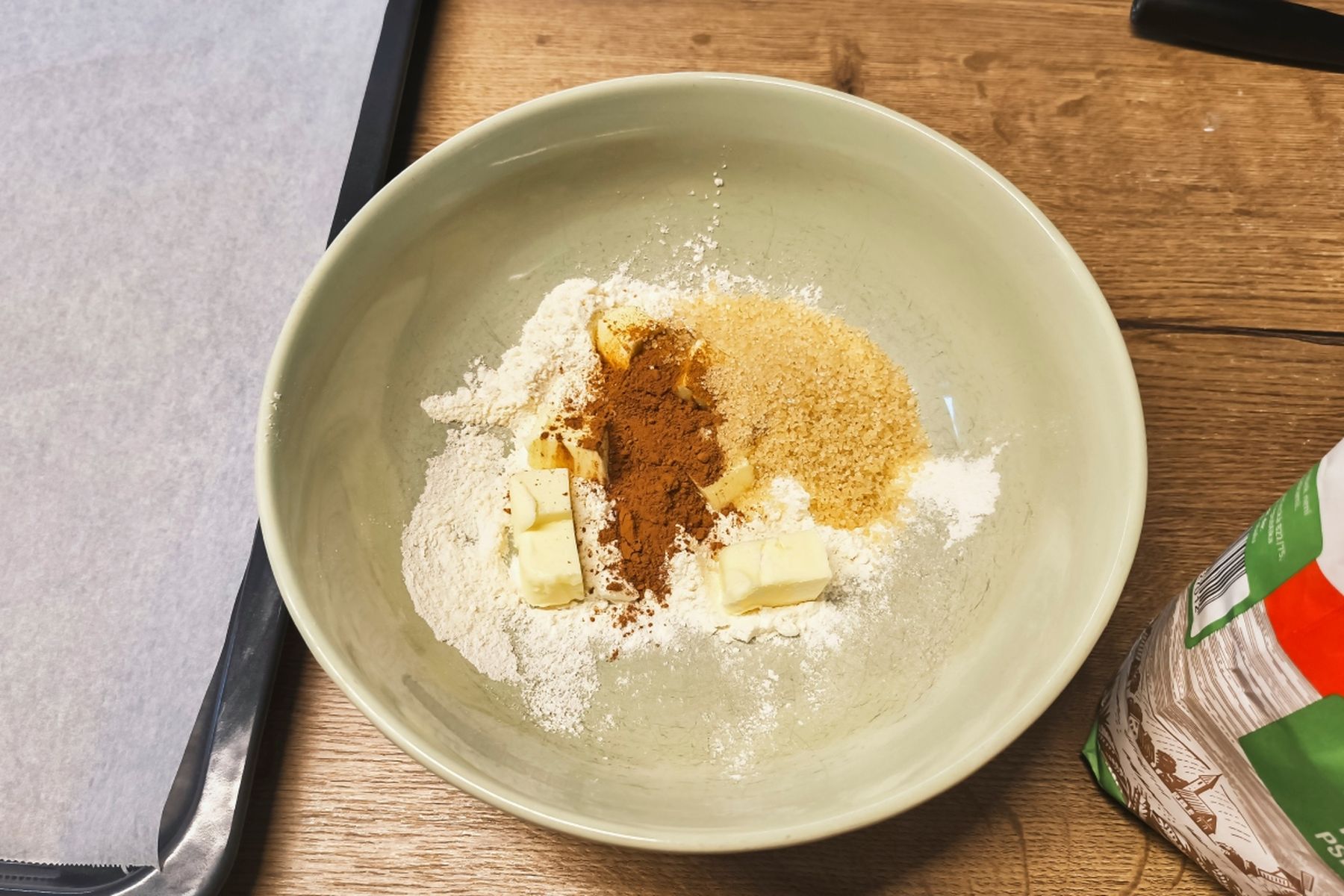 Mísa s moukou, skořicí, třtinovým cukrem a kostkami másla na dřevěném povrchu, vedle plech a pytlík s moukou. Recept připravily Ségry v kuchyni.