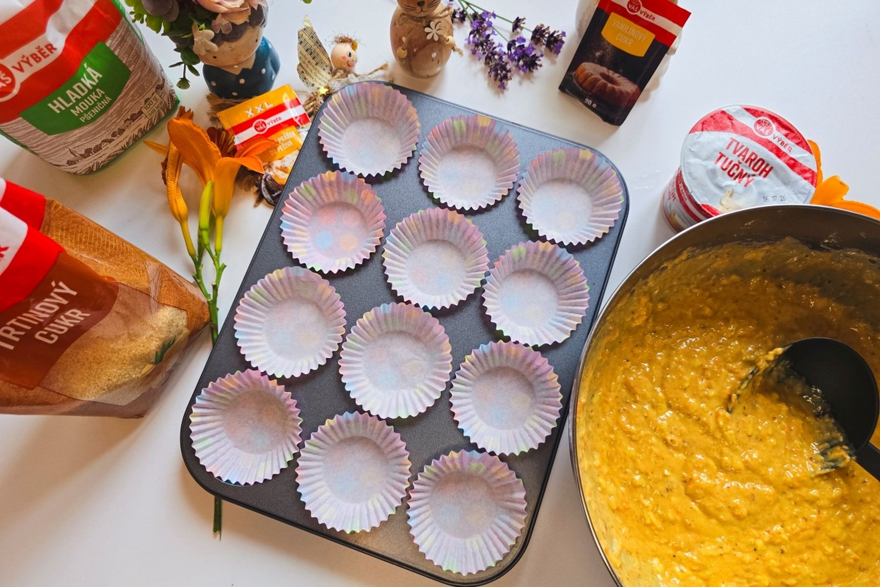 Pekařská sestava s plechem na muffiny vyloženým papírovými košíčky, surovinami jako mouka a cukr a miskou se žlutým těstem připraveným k pečení.
