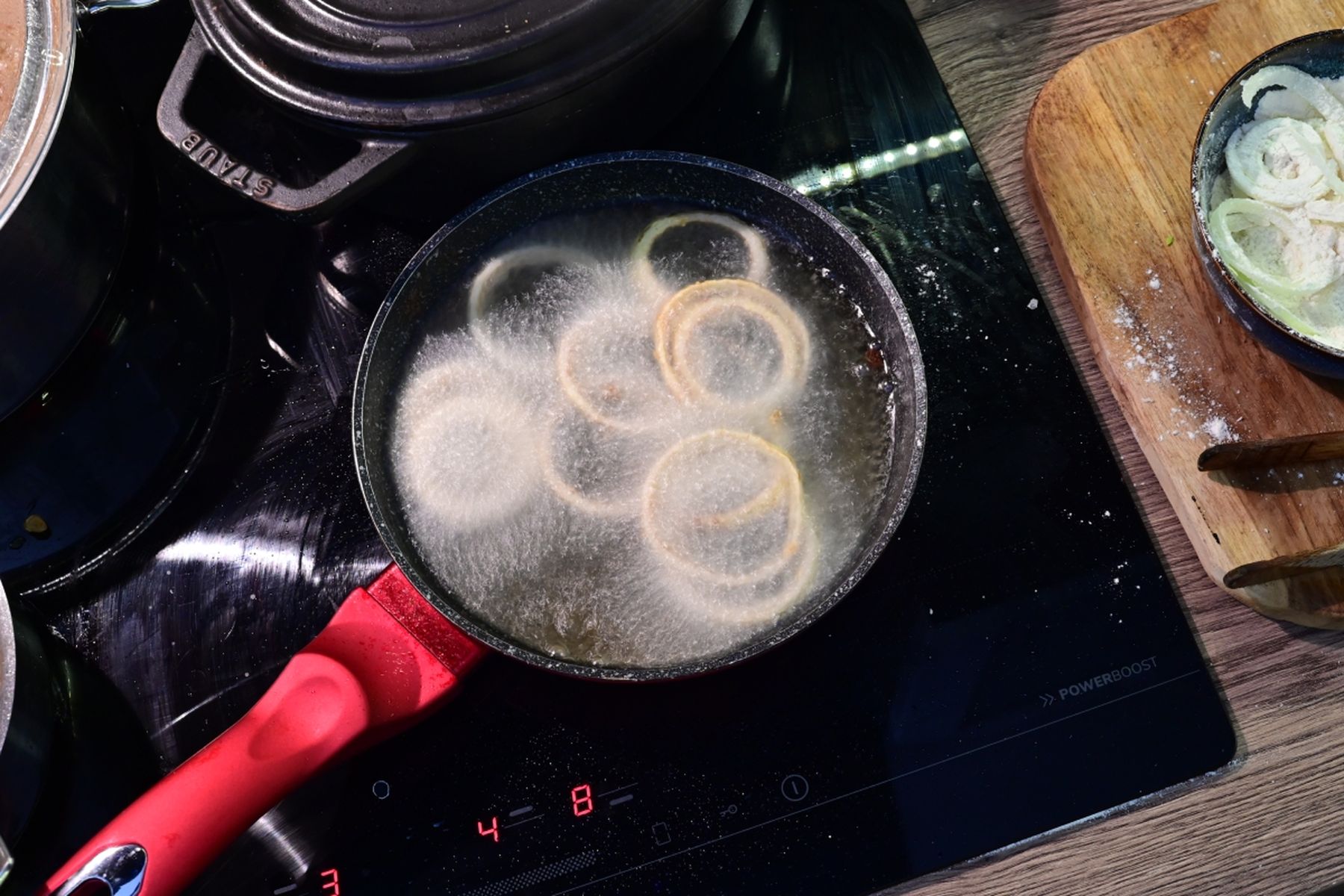 Pánev s červenou rukojetí na černé varné desce, ve které se smaží cibulové kroužky v těstíčku. Vedle leží prkénko s obalenými cibulovými plátky. Jedná se o recept z nabídky Globus.