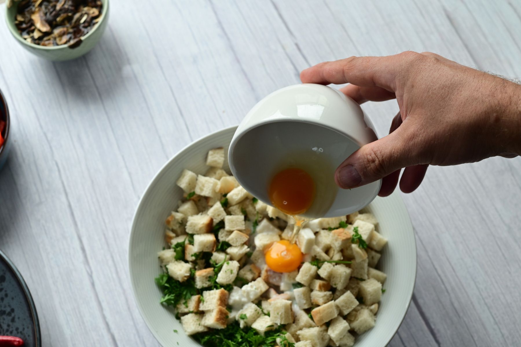 Ruka lijící vejce do misky s toustovým chlebem a bylinkami na světlém dřevěném stole, vedle mísa s dalšími surovinami. Recept na kefírový knedlík z hypermarketu Globus od Vojty Poštulky.