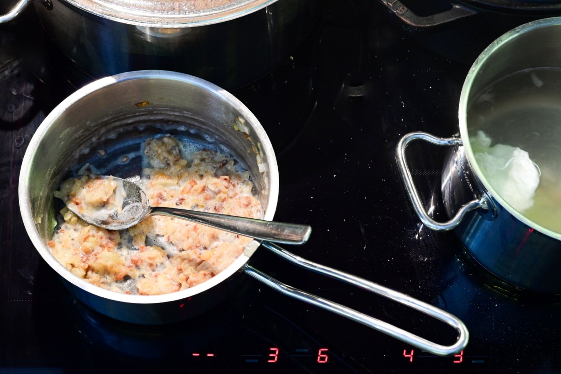 Dva nerezové rendlíky na černé varné desce – vlevo krémová směs s lžící, vpravo voda s pošírovaným vejcem. Jedná se o recept z nabídky Globus.