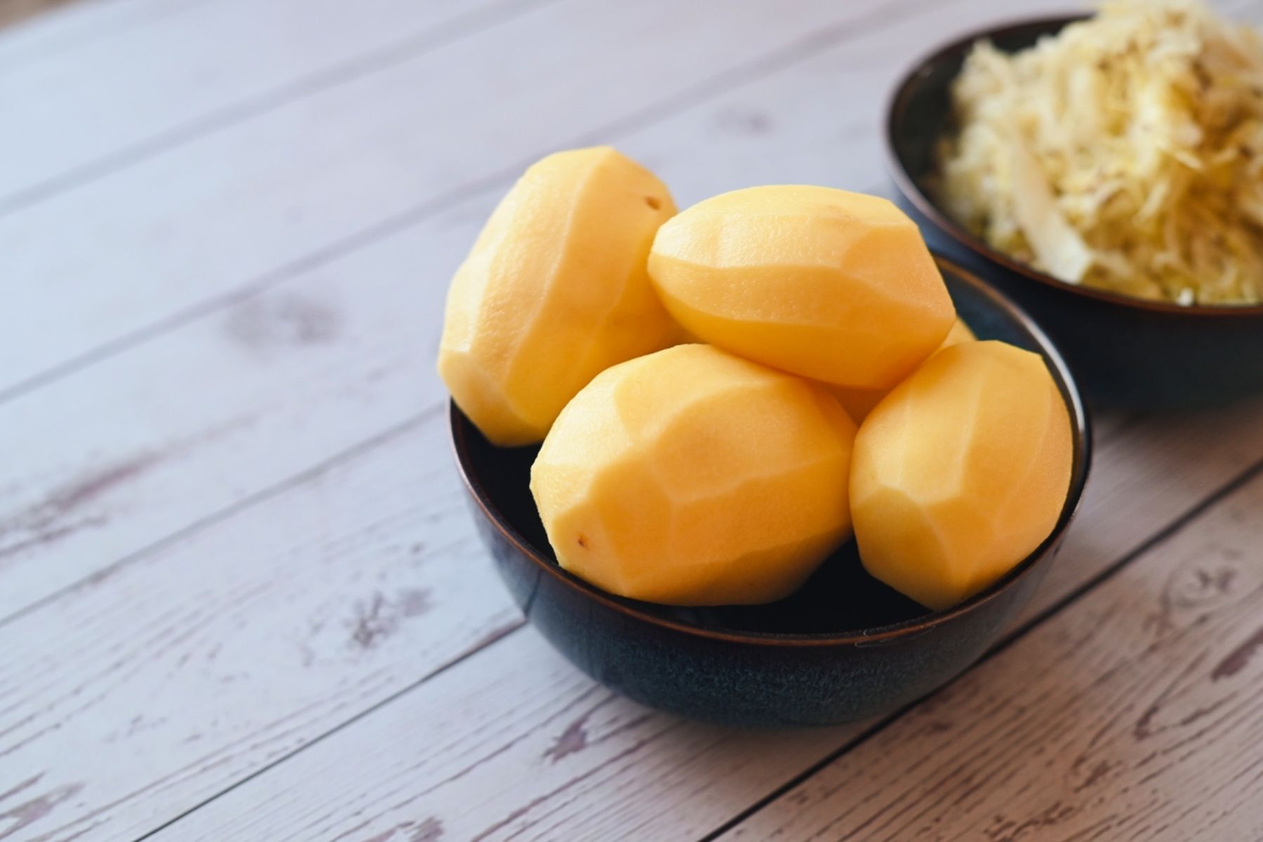 Čtyři oloupané brambory v tmavé misce na bílém dřevěném povrchu, v pozadí miska se zelím..