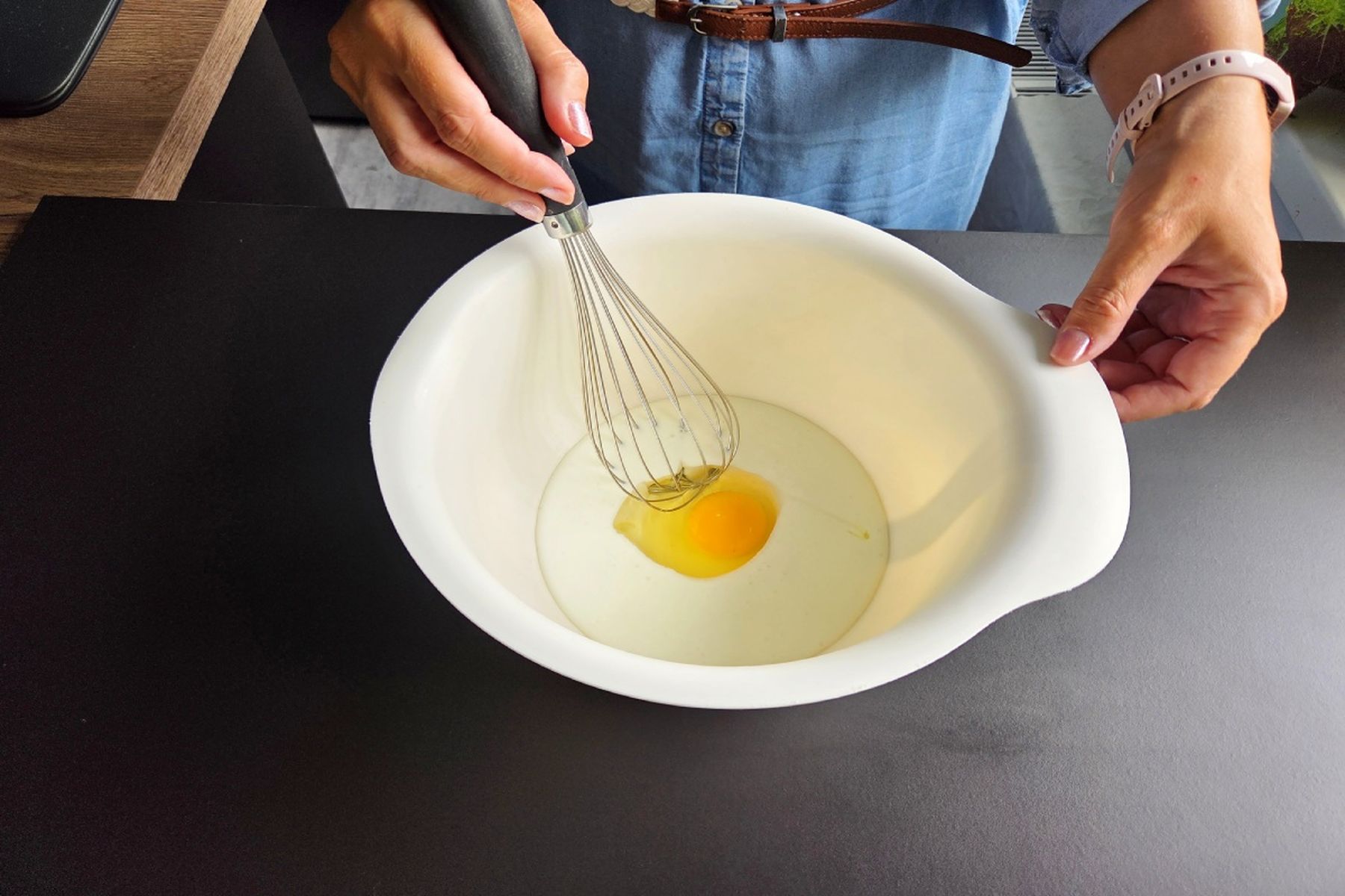 Kuchařka drží metličku nad mísou s vejcem a kefírem na tmavé pracovní desce. Recept připravily Ségry v kuchyni.