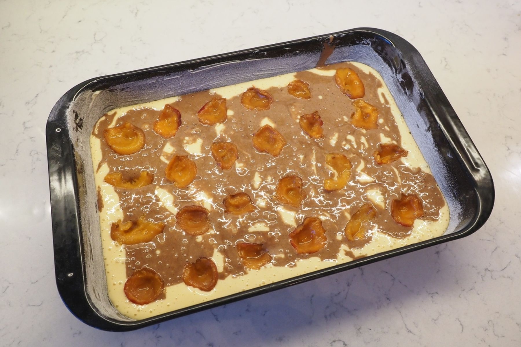 Obdélníkový pekáč s těstem na koláč, ozdobený půlkami meruněk a mramorovaným kakaovým těstem, připravený k pečení.