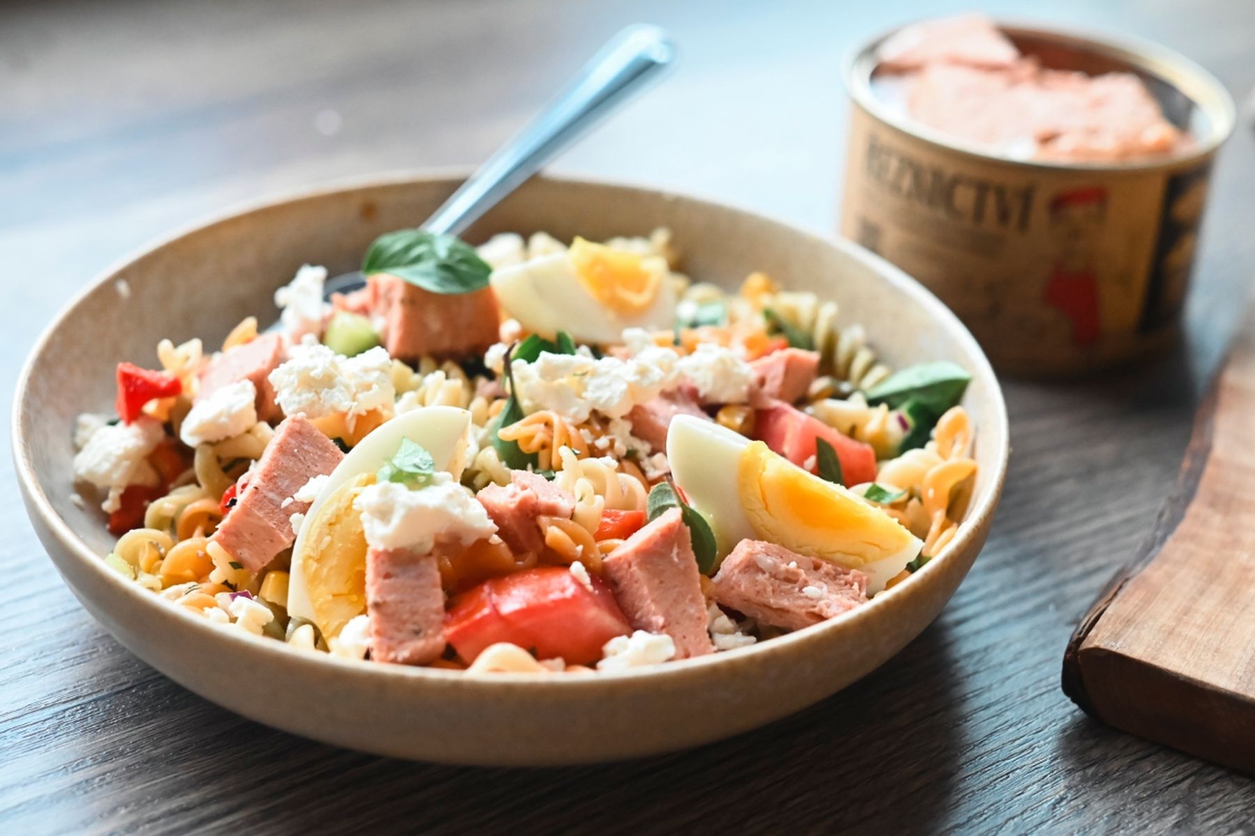 Miska těstovinového salátu se šunkou, vařenými vejci, rajčaty, špenátem a sýrem feta na dřevěném stole, v pozadí konzerva z Řeznictví Globus. Recept připravil Vojta Poštulka.